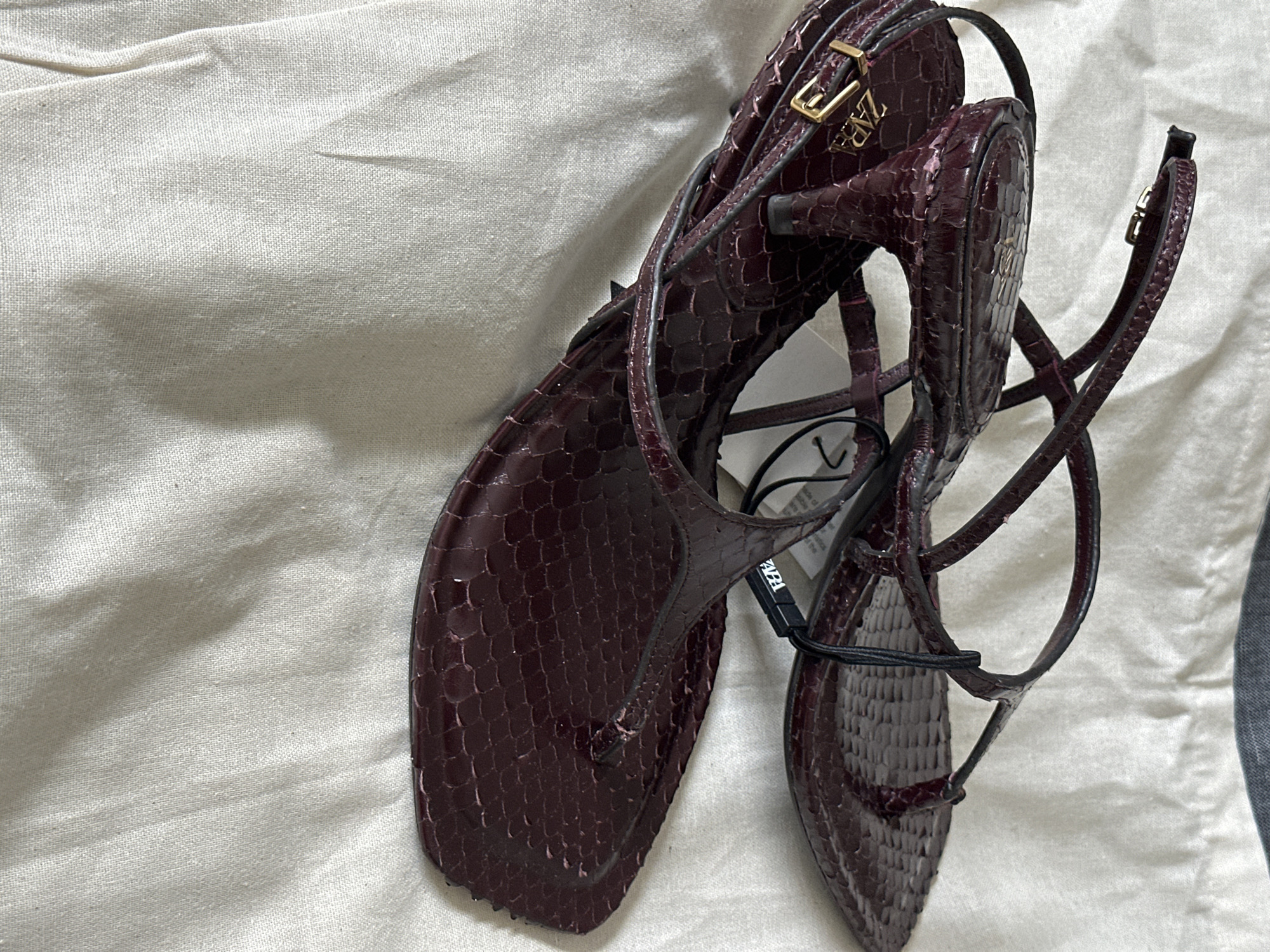 A pair of burgundy, snakeskin-textured flat sandals with thin straps and a small kitten heel are displayed on a wrinkled white fabric surface. The sandals have a thong strap between the toes and an ankle strap with a gold buckle. A white tag with black text, likely a brand tag, is attached to one of the sandals. The background is an indistinct white fabric, possibly a tablecloth or bedsheet.