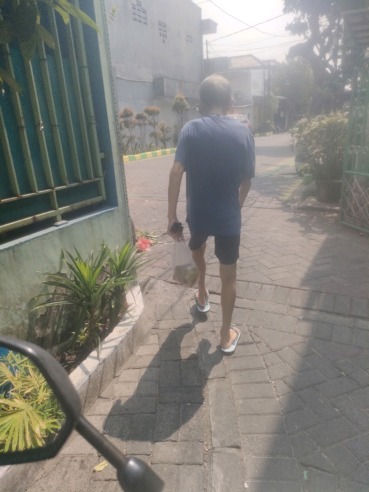 A person walks along a paved sidewalk in Surabaya, Indonesia, carrying a plastic bag. The person is viewed from behind, wearing a dark blue t-shirt and black shorts, with white and blue flip-flops. The sidewalk is constructed of grey interlocking bricks, and to the left, a concrete planter box contains green, spiky plants. A green metal fence with vertical bars runs along the side of the planter box, and beyond that, a light grey building with small, rectangular window openings is visible. The sky is bright and hazy, suggesting daytime with strong sunlight casting shadows. In the foreground, the edge of a motorcycle's black mirror is visible, reflecting green foliage. The overall environment suggests a residential or commercial area.