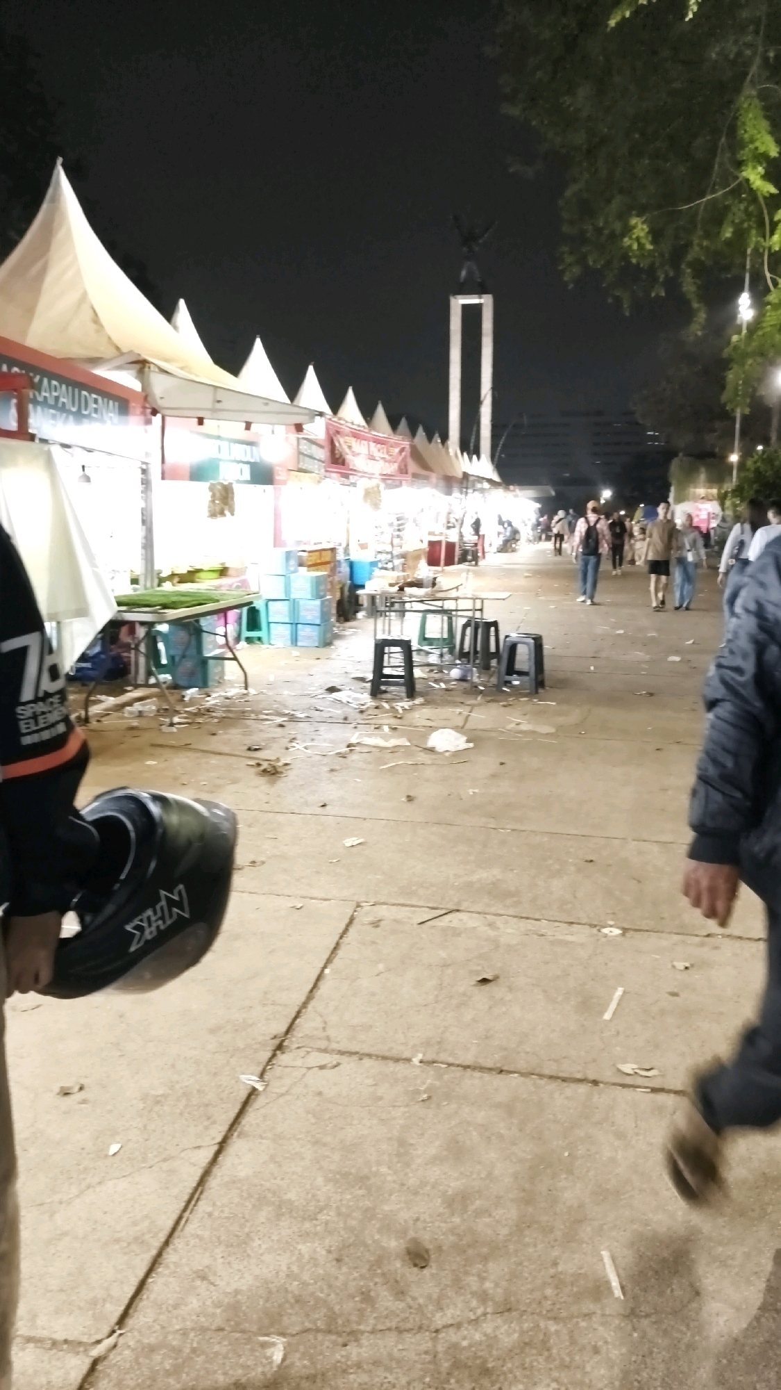 A nighttime outdoor scene depicts a bustling market with numerous stalls and a tall monument in the background. The market is lined with white, conical tents, under which vendors appear to be selling various goods. Some stalls are illuminated by artificial lights. In the middle ground, several tables and stools are set up, suggesting a seating or dining area. The ground is paved with concrete and scattered with debris, including food wrappers and plastic bags. A person on the left is holding a black object, possibly a helmet, with an "NHK" logo visible. In the distance, a large monument, likely the Monumen Nasional (Monas) in Central Jakarta, Indonesia, stands prominently against the dark sky, illuminated from below. Several people are walking through the market, some in small groups, appearing to browse or move through the area. The overall atmosphere suggests a lively evening event or marketplace.