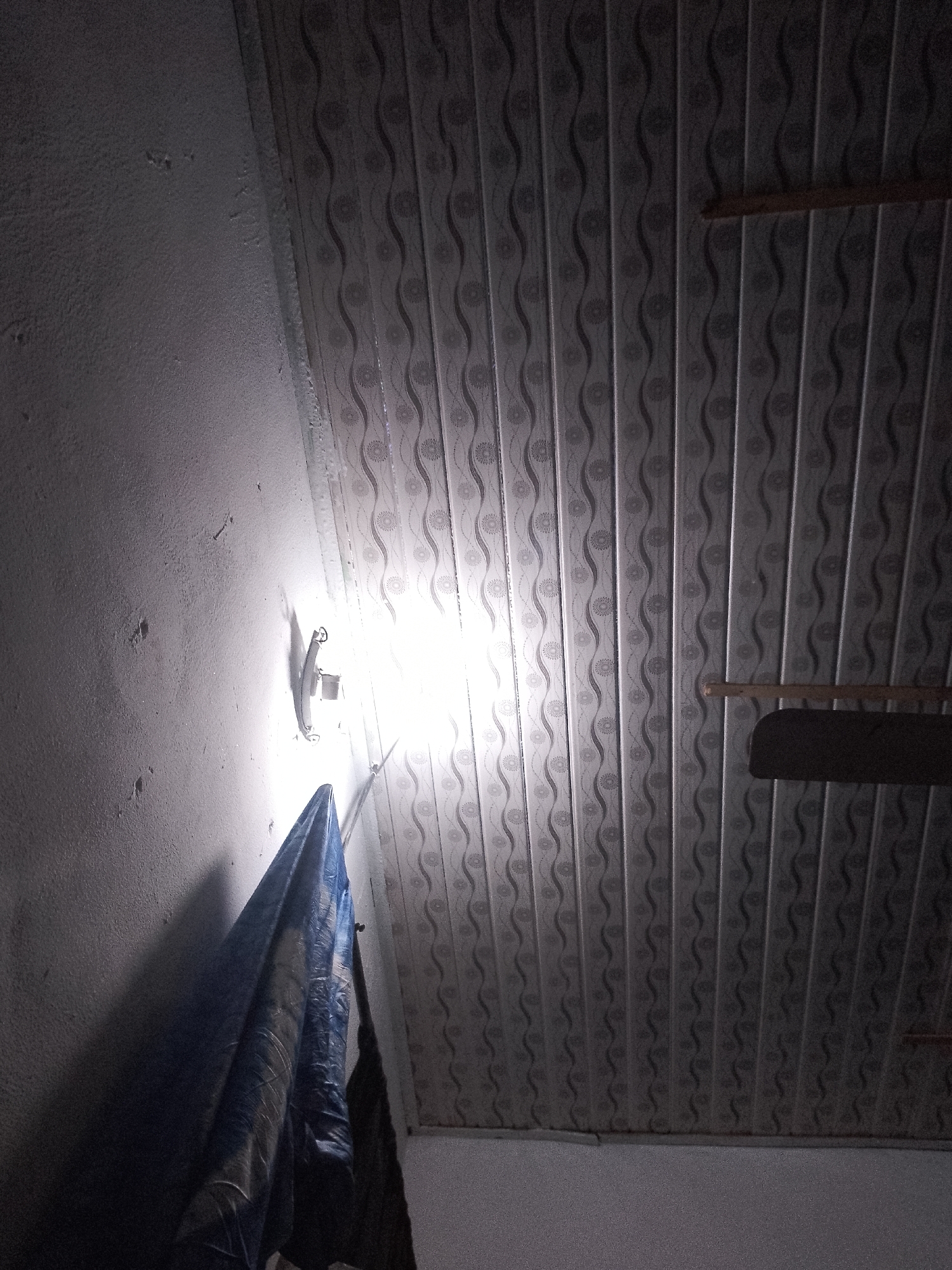 The image shows an indoor scene with a white textured wall on the left and a patterned ceiling on the right. A bright light fixture is mounted on the wall near the center of the image, casting a strong glow. A dark blue tarp or fabric hangs from the wall, partially obscuring the light. The ceiling is made of long, narrow panels with a repeating wavy pattern and circular floral motifs. A wooden support beam is visible on the ceiling, along with a dark object that appears to be part of a ceiling fan. The lighting in the scene is uneven, with the area around the light fixture being brightly illuminated and other parts of the image in shadow.