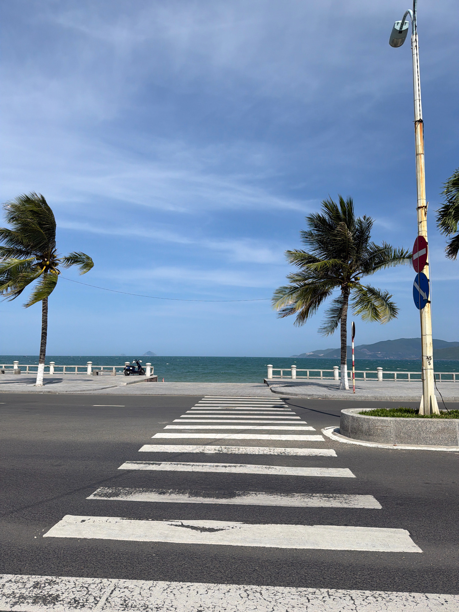 The image depicts a street scene in Nha Trang, Vietnam, facing the coast on a clear day. In the foreground, a paved road with a white pedestrian crossing is prominent. Two palm trees, one on the left and another on the right, frame the view. The tree on the left is leaning significantly to the left, its fronds appearing wind-swept. The tree on the right is straighter and has a traffic light pole with two signs next to it.
Beyond the road, a paved promenade runs parallel to the sea. A white railing separates the promenade from a body of water, which is the sea. The sea is a vibrant green-blue, with visible waves. A dark-colored scooter is parked on the promenade to the left of the image's center. In the distance, mountains are visible on the horizon, and a small island or rock formation can be seen in the sea. The sky is blue with scattered white clouds. The overall impression is a coastal urban environment with a sense of a breezy day.