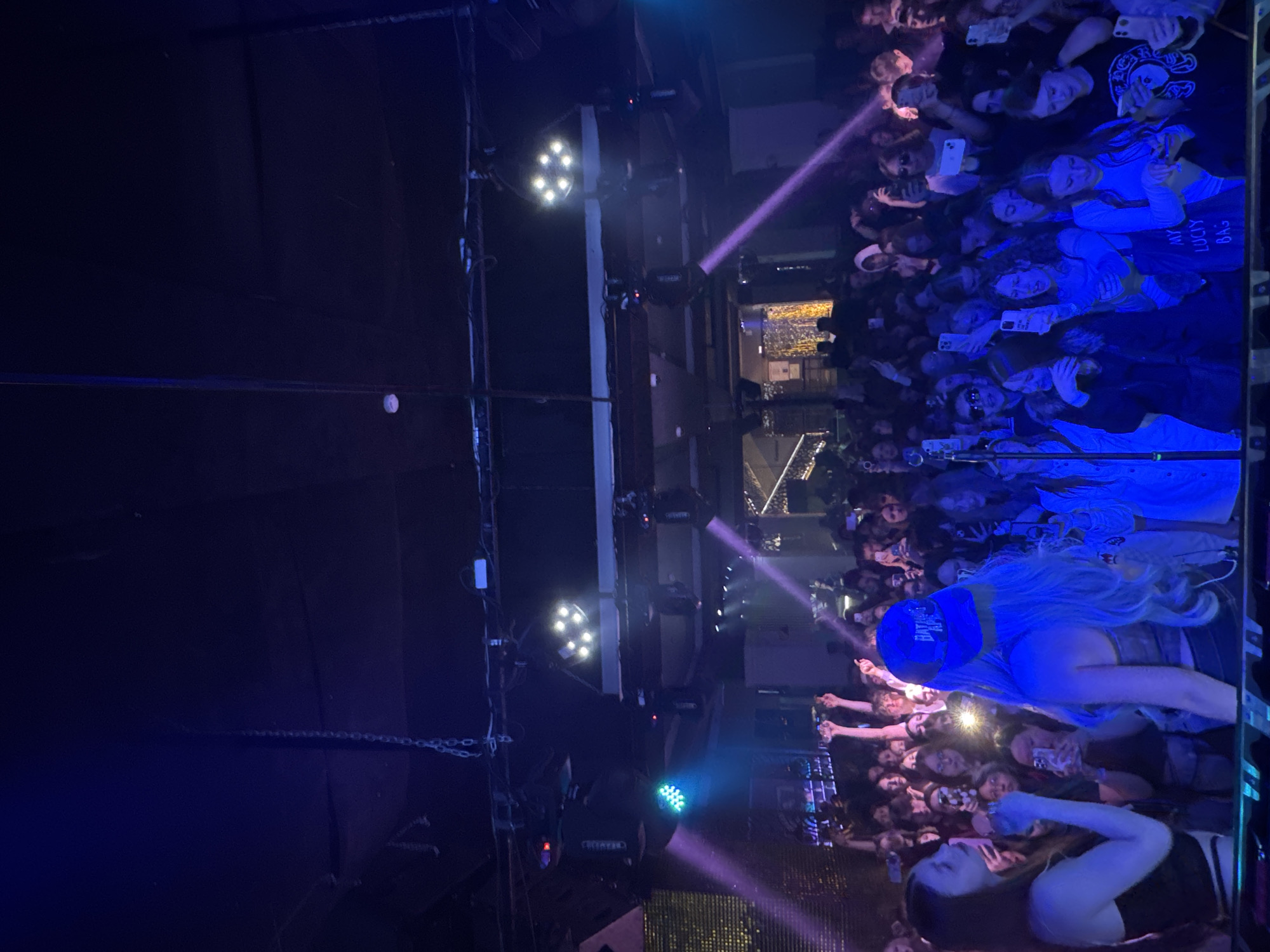 This image captures a vibrant, high-energy indoor concert or live event in Yuzhno-Sakhalinsk, Russia. The scene is dimly lit, indicative of a nighttime performance, with powerful stage lights casting purple, blue, and white hues across the venue. A large crowd of people, predominantly young adults, fills the foreground and midground, looking towards an unseen stage to the left. Many individuals are holding up smartphones, actively recording the performance or taking photos, with some flashlights from phones also visible.
The audience appears engaged and excited, with several faces visible expressing enjoyment. One person in the crowd wears a blue baseball cap backward, displaying the text "BATTLE". Another individual holds a light-colored tote bag with "MY LUCKY BAG" printed on it. Overhead, various lighting fixtures are mounted on trusses, including clusters of bright white spotlights and a rectangular light emitting a blue-green glow. Purple laser-like beams cut through the air, adding to the dynamic atmosphere. The perspective is from a slightly elevated position, overlooking the packed crowd.