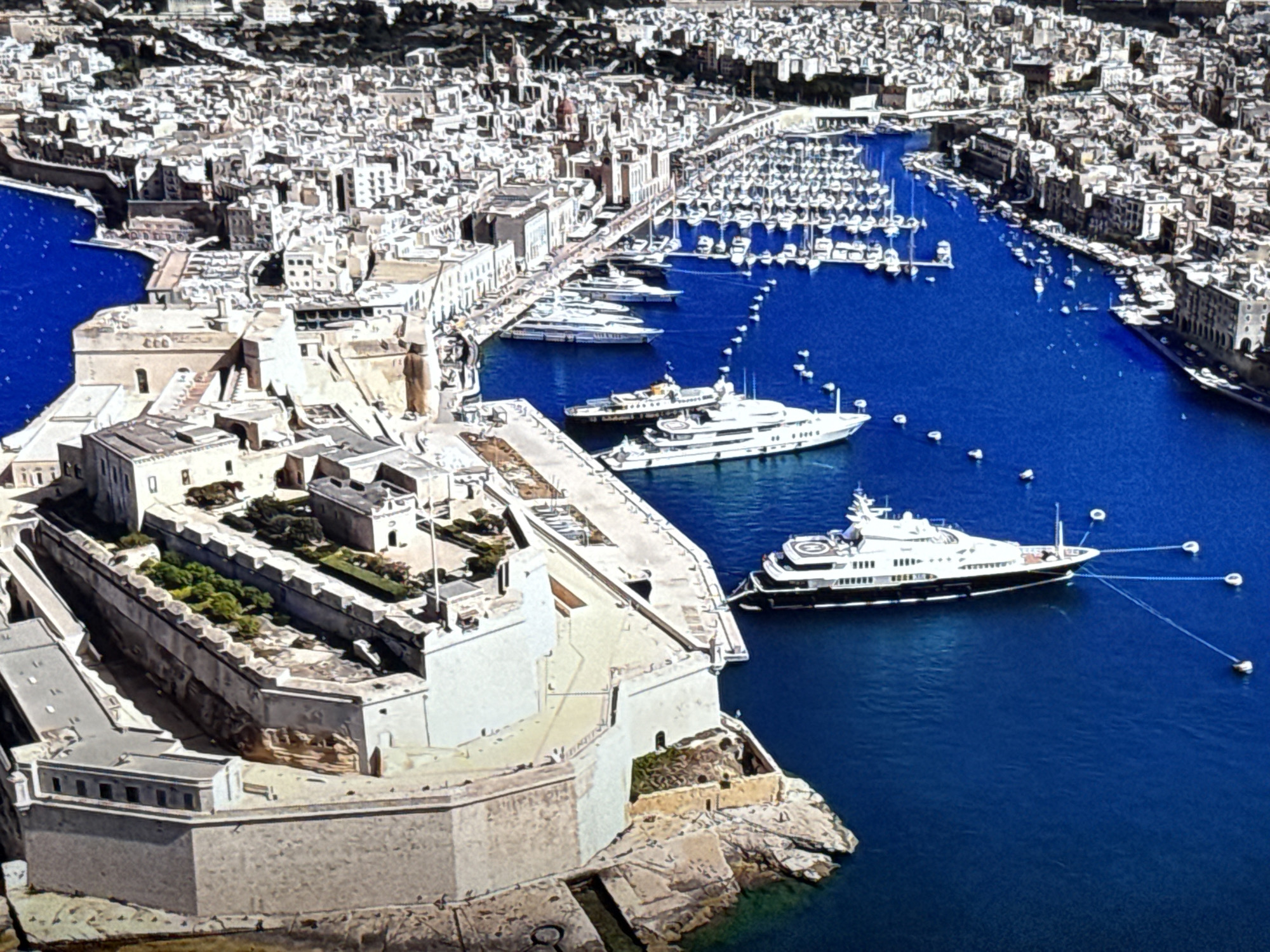 The image is an aerial view of a fortified city on a body of water. The city is densely built with white and light-colored buildings, many of which appear to be historical. The body of water is a deep blue and is filled with numerous boats, including a large luxury yacht docked near the fortifications. The fortifications themselves are massive stone structures with ramparts and battlements, extending into the water. There are also many smaller buildings and structures within the fortified areas, including what appear to be gardens. The overall impression is one of a historic port city with a strong defensive past, currently a bustling hub for maritime activity. This is a scene of the city of Valetta, Malta, not Burgas, Bulgaria.