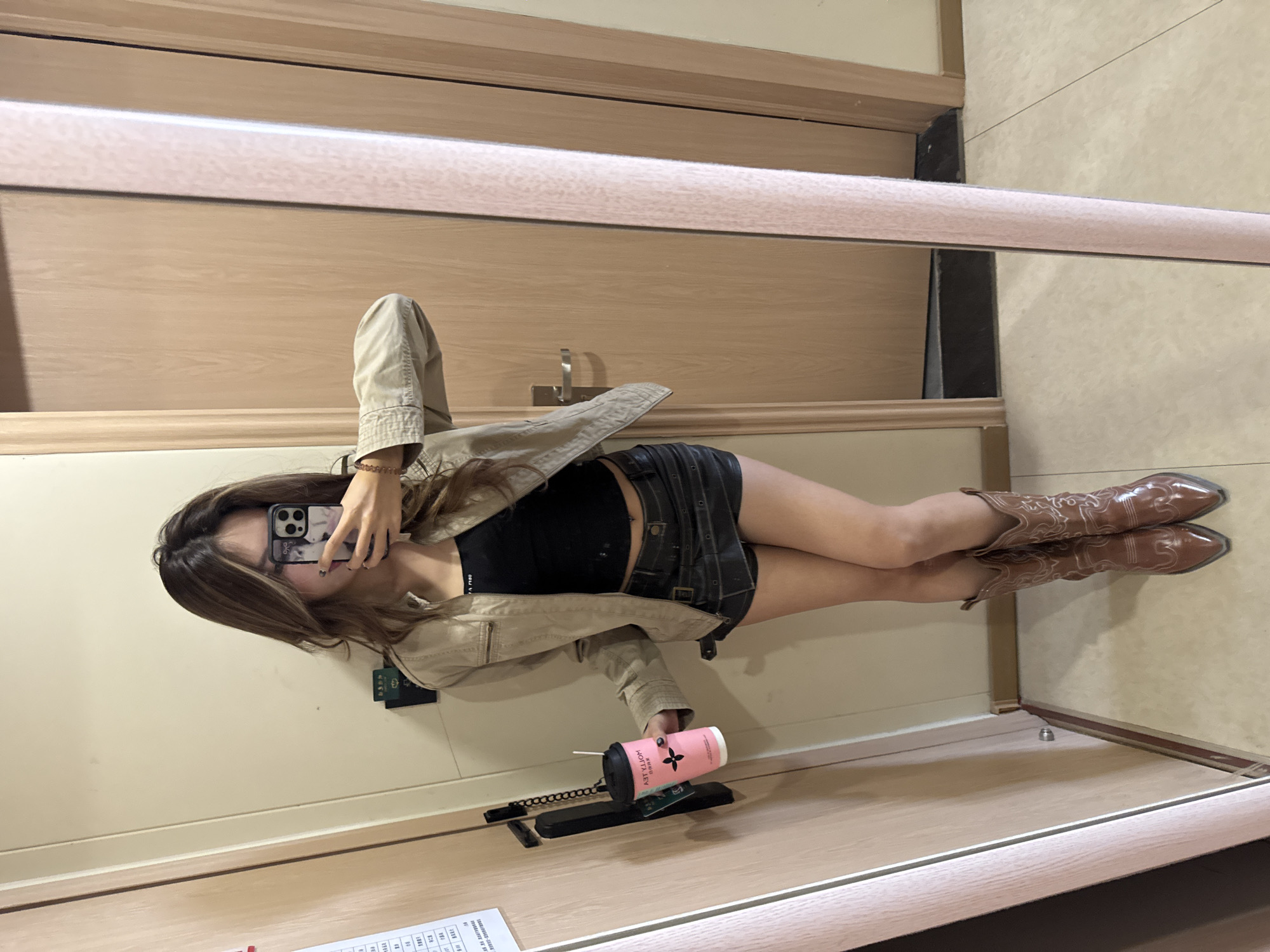 The image shows a person taking a mirror selfie in a room. The person is wearing a black top, a denim skirt, a beige jacket, and brown cowboy boots. The phone in the person's hand is used to take the photo. A pink cup and some papers are on the floor. The room appears to be in a hotel, with a light-colored wooden door and trim. The location is Dunhuang City, China, based on the papers on the floor in front of the subject.