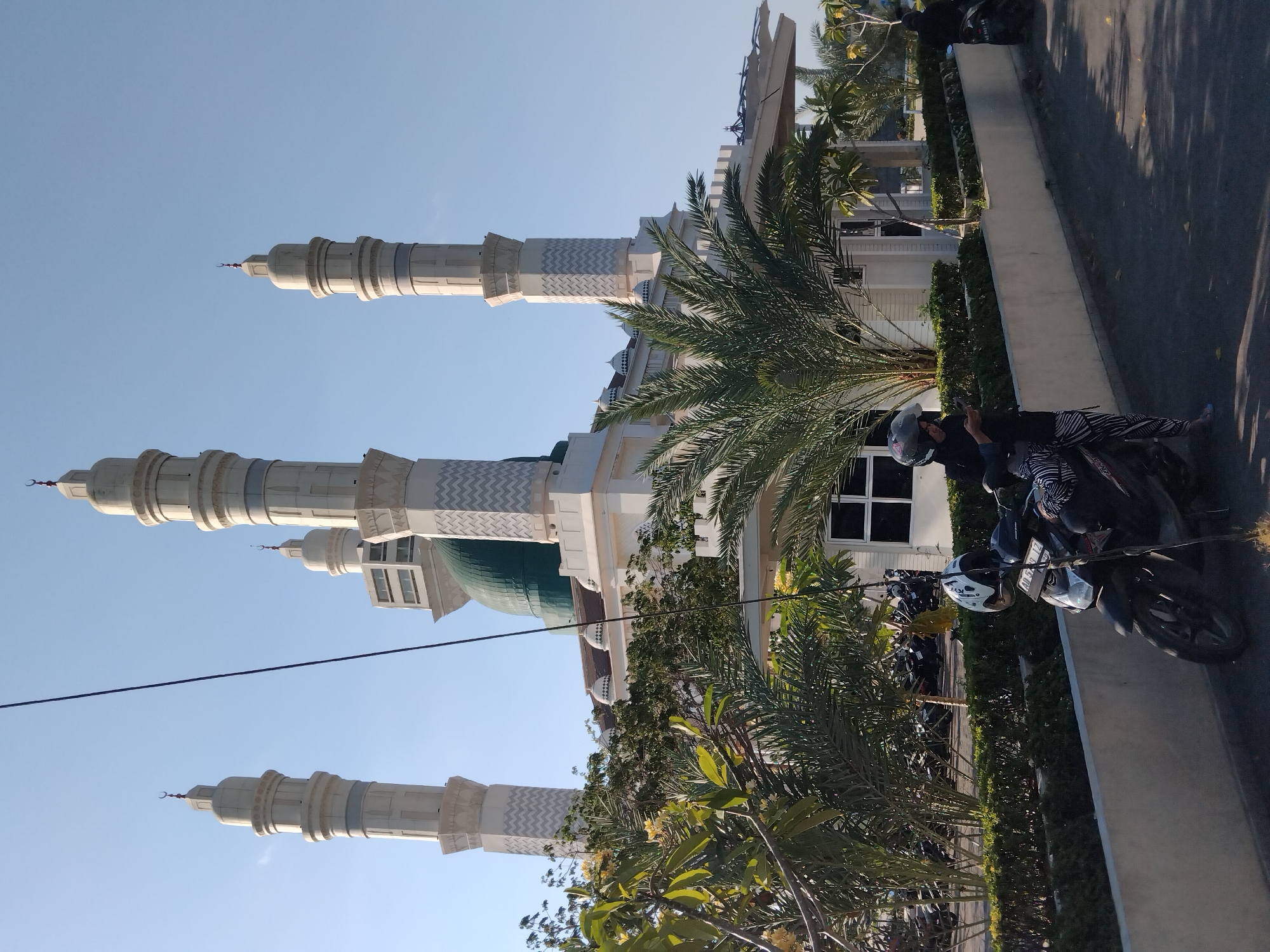 In the city of Karanganyar, Indonesia, a modern mosque with white minarets topped with crescent moons and a green dome stands under a clear blue sky. The mosque features a white facade with decorative zigzag patterns. In the foreground, a person wearing a black headscarf and patterned pants stands beside a parked motorcycle, looking at their phone. Another motorcycle is parked nearby, and a third person on a motorcycle is visible further in the background. Lush green trees and palm trees surround the mosque, with manicured hedges lining a paved walkway. The overall scene suggests a sunny day in a well-maintained urban environment.
