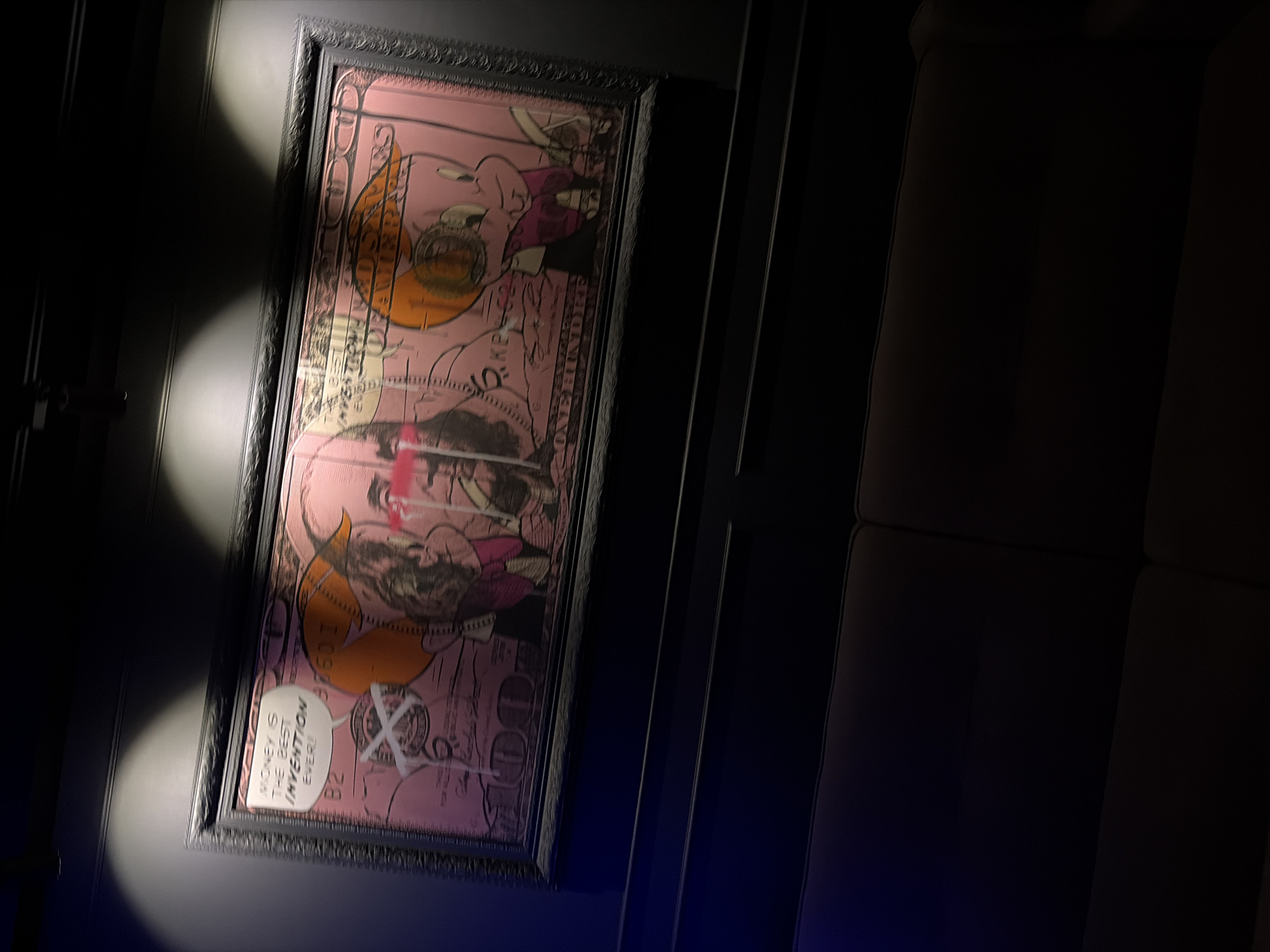 The image depicts an indoor scene focused on a framed piece of pop art illuminated by two overhead spotlights. The artwork, presented horizontally in a dark, ornate frame, is a pink-hued rendition of a United States $100 bill. The central figure, typically Benjamin Franklin, is depicted with red streaks or "tears" beneath his eyes, and white graffiti-like marks obscure parts of his face. A white speech bubble originating from his position reads, "MONEY IS THE BEST INVENTION EVER!". To the right, where Independence Hall would typically be, a cartoon pig-like character wearing an orange hat and purple clothing is depicted, also with some white graffiti marks. The background of the artwork retains faint details reminiscent of currency, including partially visible text such as "ONE HUNDRED DOLLARS" at the bottom.
The artwork hangs on a dark, possibly matte black or very dark grey, wall. Below the artwork, a dark-colored, two-cushion upholstered couch is visible, suggesting a seating area. A dark wainscoting or decorative trim runs horizontally along the wall between the artwork and the couch. The lighting in the room is dim, with the spotlights specifically highlighting the artwork, creating distinct conical beams of light on the wall above the frame. There are no identifiable visual markers within the image to determine its specific geographical location within Chaikovsky, Russia, or any other city.