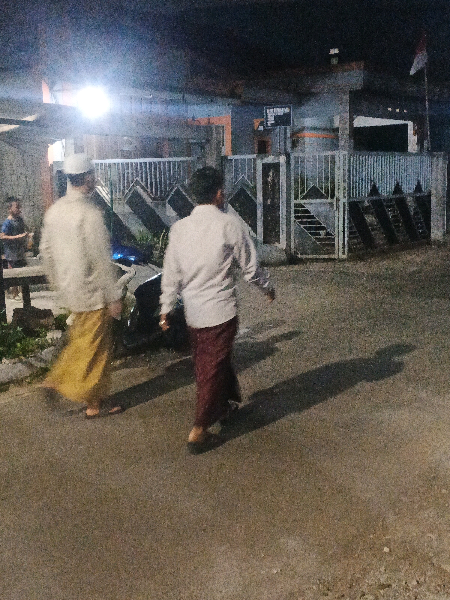 In the city of Kadu, Indonesia, at night, two men are walking away from the camera on a paved road. The man on the left is wearing a white cap, a light-colored shirt, and a yellow sarong. The man on the right is wearing a white shirt and a dark red sarong. In the background, there is a building with a gate and fencing. A bright light illuminates the scene from above and to the left. To the left of the road, there is some vegetation and a motorcycle. Another person in a blue shirt is visible in the background on the left. The shadows of the two main subjects are cast on the road.