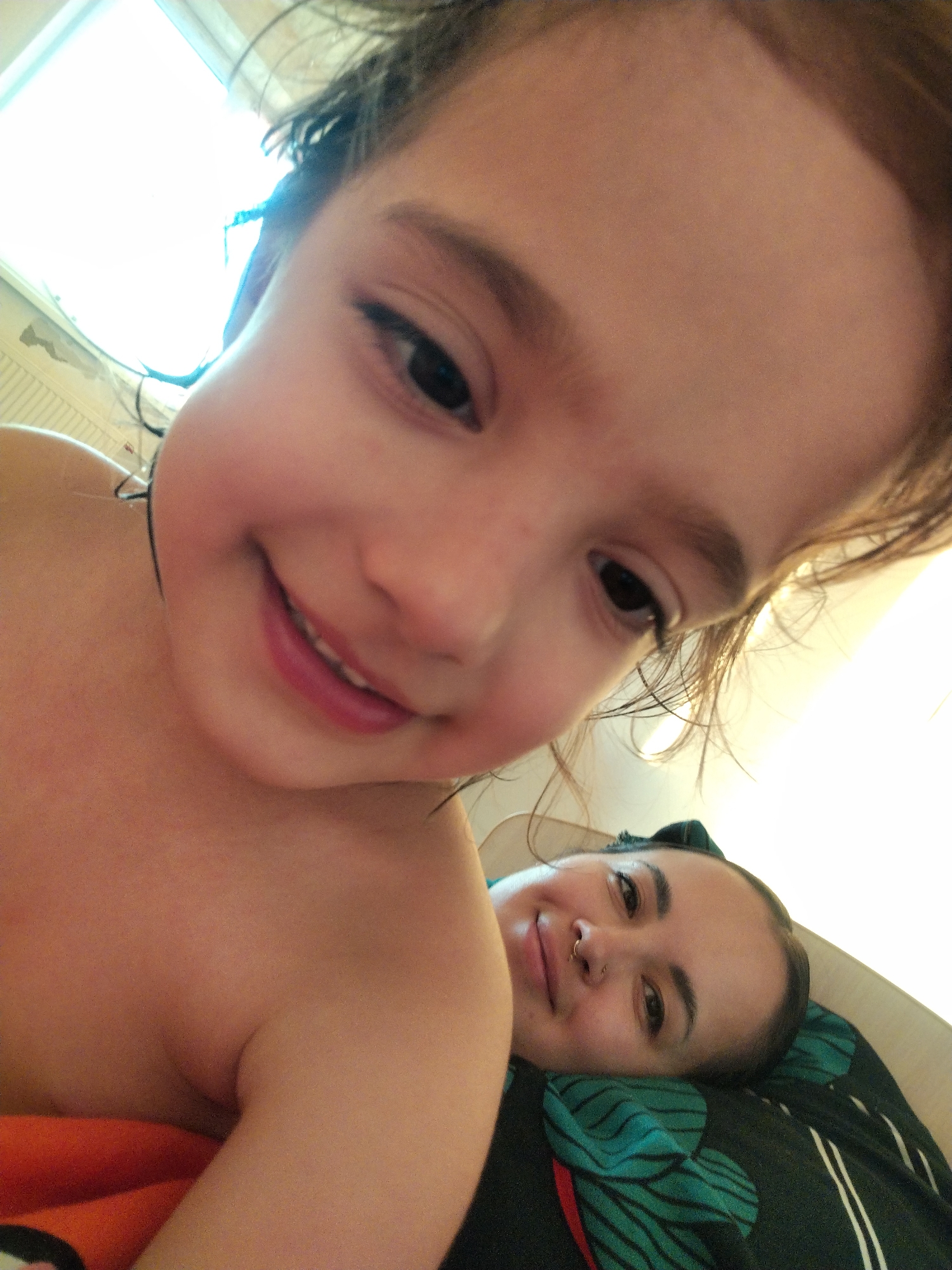 The image shows a close-up, selfie-style shot of a young child and an adult woman. The child is in the foreground, with a broad smile, positioned higher and closer to the camera. The woman is in the background, reclining on what appears to be a bed or pillow, also smiling slightly. The light source is from above, suggesting a window. The overall setting appears to be indoors. The location in the city of Noviy, Russia cannot be determined from the provided image.