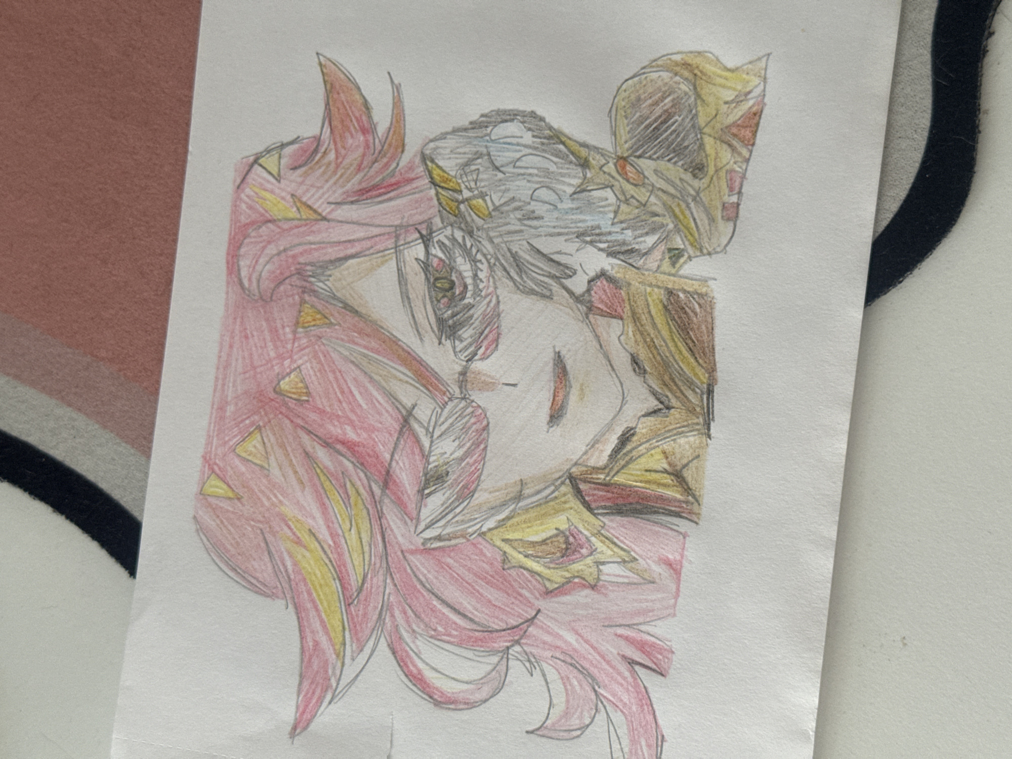 The image shows a colored pencil drawing of a character with red and yellow hair, wearing a headpiece that is colored in shades of gray, gold, and brown. The drawing is on a white sheet of paper, which is placed on a surface that has a mix of patterns and textures. Part of a rug with brown, beige, and black stripes is visible in the background on the left side of the drawing. On the right, a white surface with a black outline can be seen. The location is not indicated within the image.