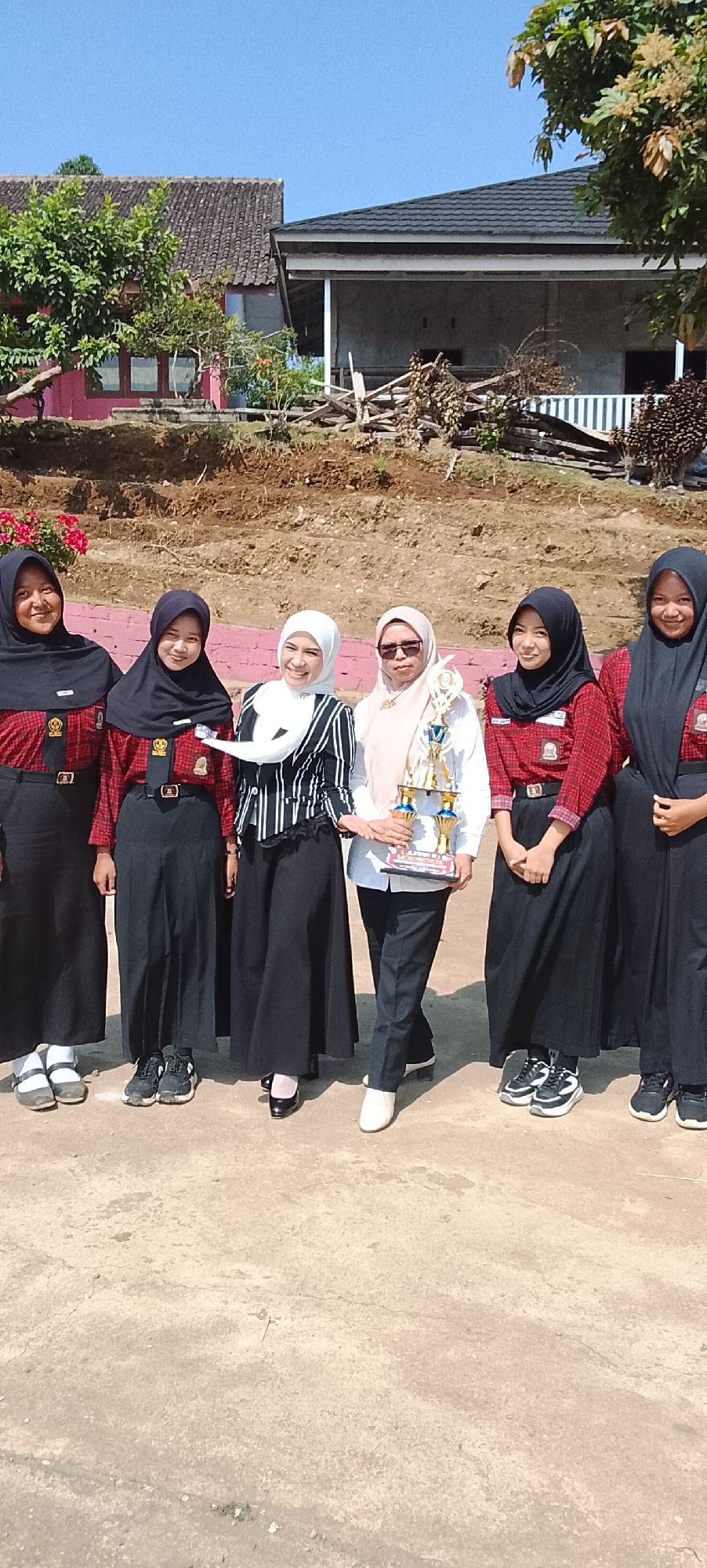 Six individuals, appearing to be students and a teacher, pose for a photograph outdoors. The five individuals on the left and right are dressed in similar school uniforms consisting of red and black checkered long-sleeved shirts with black ties and emblem pins, paired with long black skirts. All are wearing headscarves, with five in black and one in a lighter color. The central individual, presumably a teacher, wears a white shirt and a black and white striped jacket, also with a headscarf. This individual holds a golden trophy with blue accents. They are standing on a cracked concrete ground, with a reddish-brown dirt embankment and some greenery behind them. In the background, there are buildings with tiled roofs and wooden structures. The lighting suggests it is daytime with clear skies. The exact location within Jepara, Indonesia, is not specified by visual cues in the image.