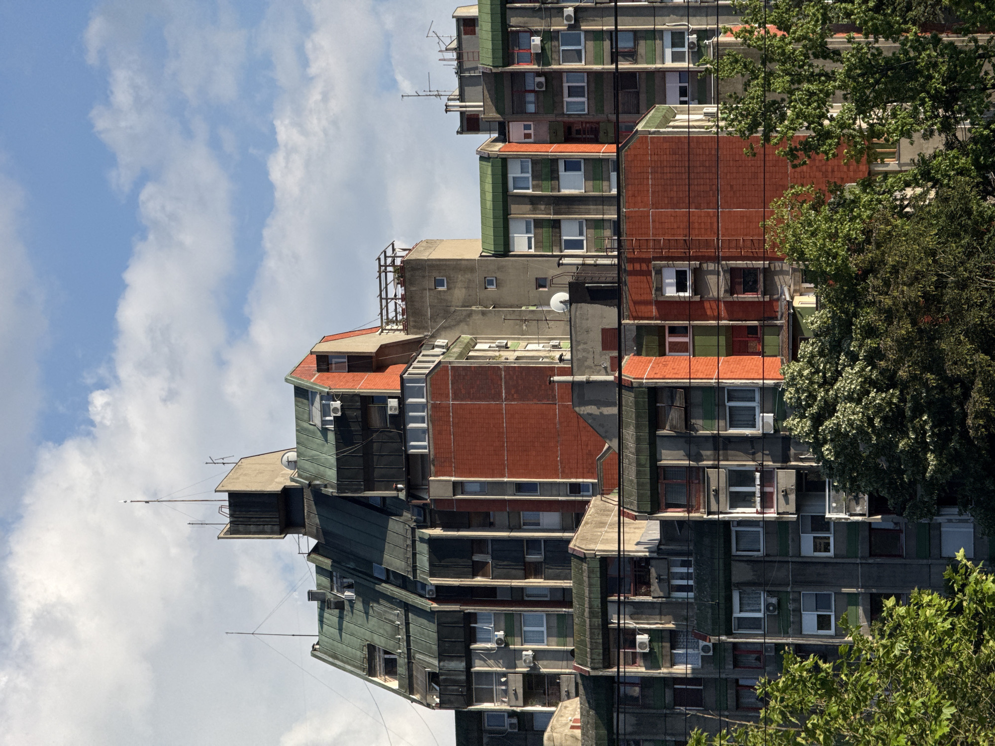 The image shows an exterior view of a multi-story apartment building in the city of Vozdovac Urban Municipality, Serbia. The building is constructed with a mix of concrete and brick, and features a unique, stacked design. The structure appears to have multiple sections, some with green accents and others with red brick. Numerous windows and air conditioning units are visible on the exterior walls. The sky is blue with some white clouds. Trees with green foliage are visible in the foreground. The image is oriented vertically, suggesting it was taken from a street-level perspective or has been rotated.