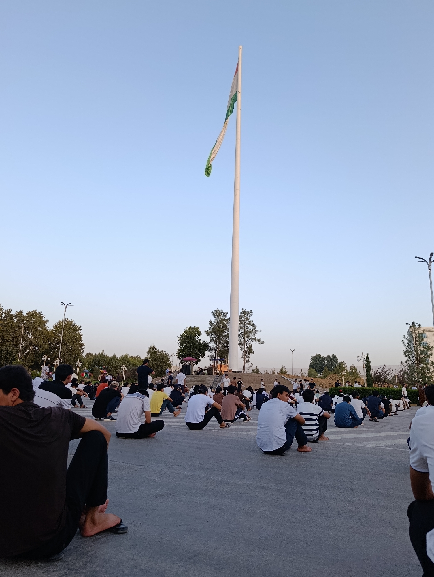 In Danghara, Tajikistan, a gathering of people is observed in an open, paved area, possibly a plaza or park. A tall flagpole with a flag is prominently positioned in the background. The flag appears to be white, green, and red. A large group of individuals, predominantly young men and boys, are seated on the ground, facing various directions. Many are dressed in casual attire, including t-shirts, long-sleeved shirts, and pants. Some are barefoot, while others wear sandals or shoes. The overall atmosphere suggests a community event or a moment of collective assembly. The surrounding environment includes trees and what appears to be a building or structure in the distance. The sky is clear and light blue, indicating daytime, possibly late afternoon or early evening due to the soft lighting.