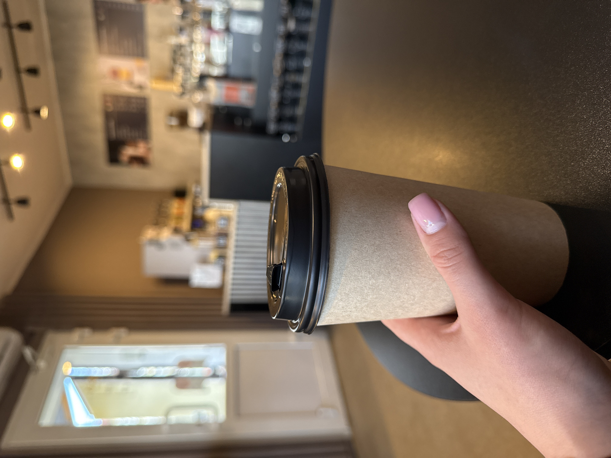 A tan paper coffee cup with a black plastic lid is held in a person's right hand. The hand has pale skin and a light pink nail polish. The cup is resting on a black surface that appears to be a table. The background shows the interior of a coffee shop, including a window, counter, and shelves. The location is the city of Chaikovsky, Russia.