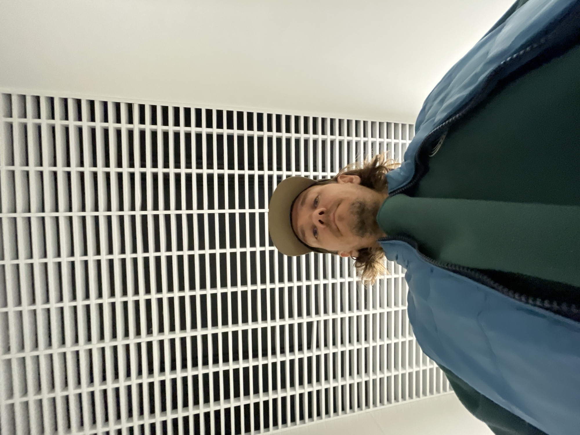 The image features a man positioned sideways against a white background dominated by a prominent white grid pattern. The man occupies the right and lower central portion of the frame. He has medium-length brown hair, light stubble or a short beard, and is wearing a light brown baseball cap. His face is turned upwards, looking towards the top edge of the image. He is dressed in a dark green long-sleeved top and a light blue puffy jacket, worn open.
The background consists of a large, uniformly white grid structure, likely a ceiling vent, air diffuser, or decorative wall panel, which covers the left two-thirds of the image. The grid is composed of numerous vertical and horizontal white bars, creating a visually striking pattern with dark spaces between the bars. Above the grid and to the right of the man's head is a plain white surface, presumably a ceiling or wall.
The activity appears to be a low-angle photograph or selfie taken indoors, with the subject looking directly at the camera. The man's expression is neutral and calm. The lighting is bright and even, characteristic of indoor artificial light. There is no visible text in the image.