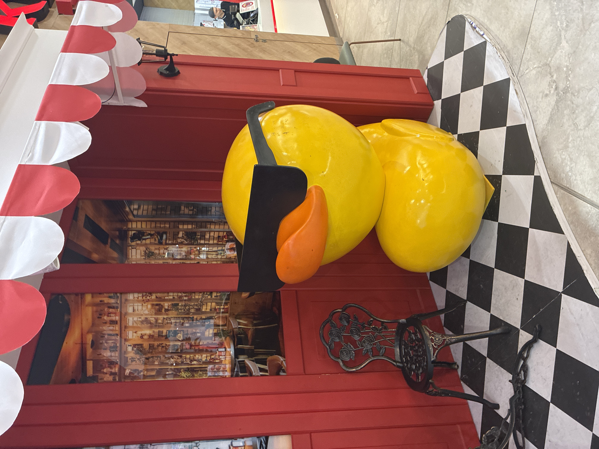 The image depicts a large, yellow, stylized duck statue wearing oversized black sunglasses and an orange beak. The statue is positioned in front of a bright red structure with a red and white striped awning. To the left of the duck is a small, ornate black metal chair with a rose design. The ground is covered in a black and white checkered tile pattern. In the background, through a windowed section of the red structure, shelves stocked with various items are visible, resembling a display in a shop or restaurant. The overall scene appears to be outdoors or in a public indoor space, possibly a mall or a themed area. The location is identified as Sofia, Bulgaria.