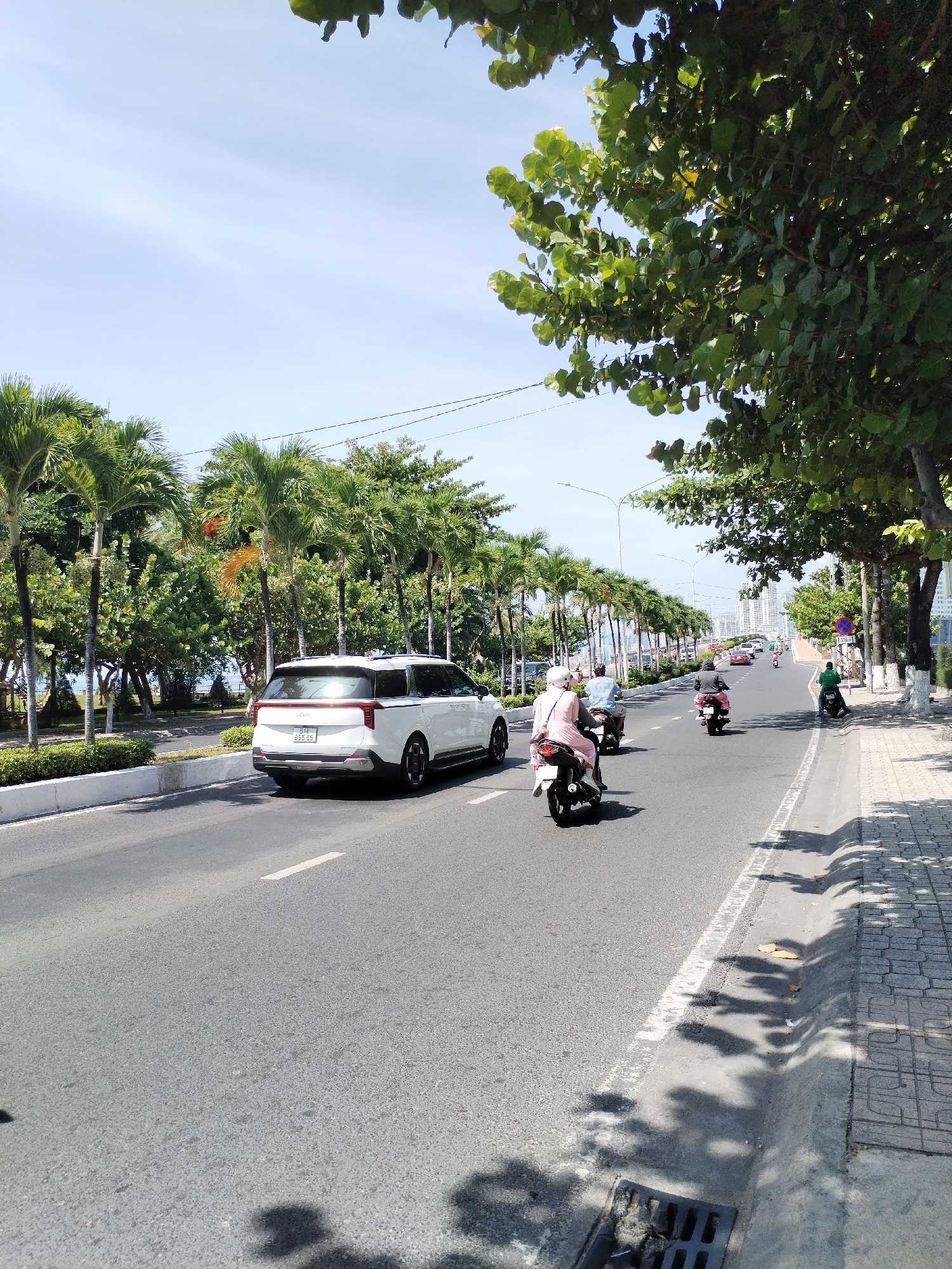 The image depicts a sunny day on a multi-lane road in Nha Trang Ward, Vietnam. A white SUV is parked on the right side of the road, and several motorbikes are in motion. On the left side of the road, there is a line of palm trees and other lush green vegetation. In the distance, tall buildings are visible, suggesting an urban setting. The road is paved and has clear lane markings. The sidewalk on the right is paved with tiles and shaded by trees. The sky is bright blue with some scattered clouds.