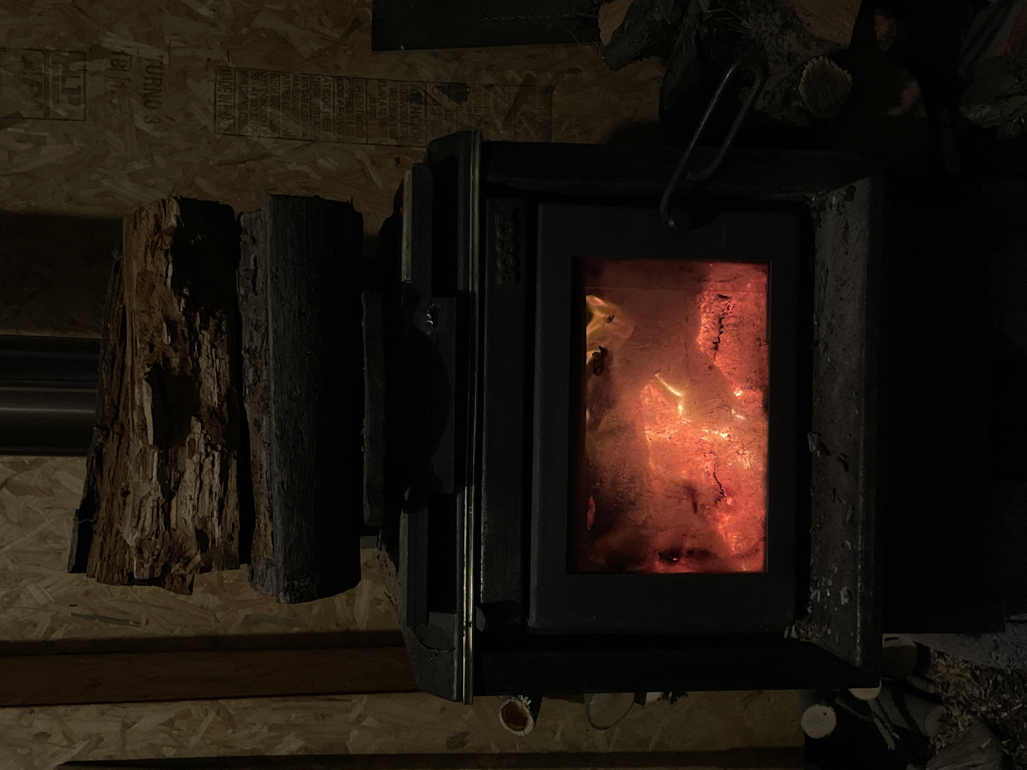 A wood-burning stove is depicted in a dark room. Flames are visible through the glass door of the stove. Two pieces of firewood are positioned next to the stove on the left. A pile of firewood is stacked atop the stove on the right. The wall behind the stove is constructed of plywood. There are written labels on the plywood. The OCR provides some information: "POSICK 525", "ATENCION", "FIJAR A 10 MM DEL BORDE", "MADE IN BRAZE", "TURNO 3". The environment appears rustic. There is no evidence to suggest the location is in Ushuaia, Argentina.