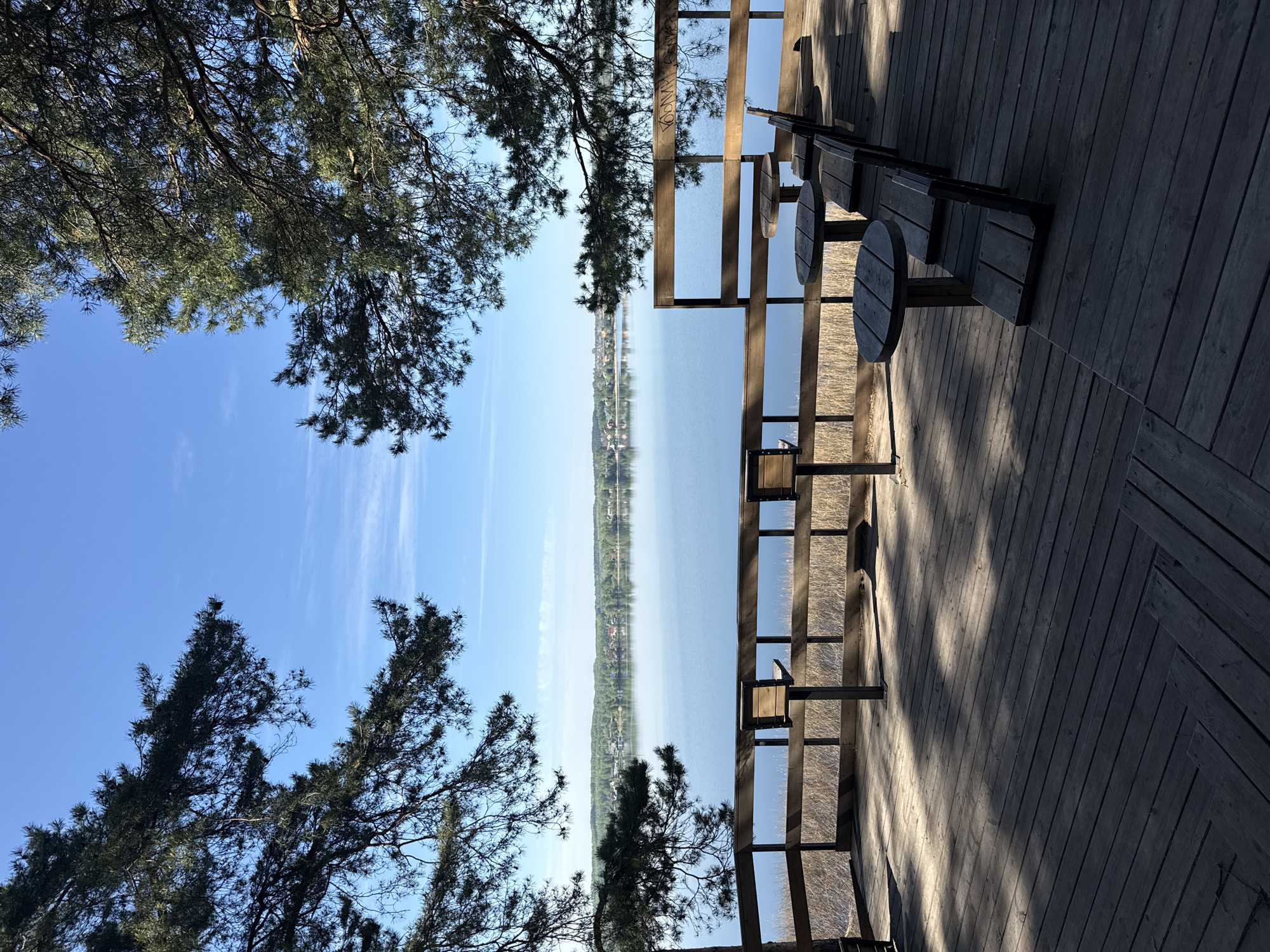 The image depicts a scenic view from a wooden deck or platform overlooking a body of water, likely the Votkinsk Reservoir in Votkinsk, Russia. The deck has a wooden railing and features benches and small round tables. Trees partially frame the scene on the left side of the frame. The body of water stretches towards the horizon, with a distant shoreline visible. The sky is blue with some wispy clouds. The overall perspective is rotated. Sunlight casts shadows across the wooden deck.