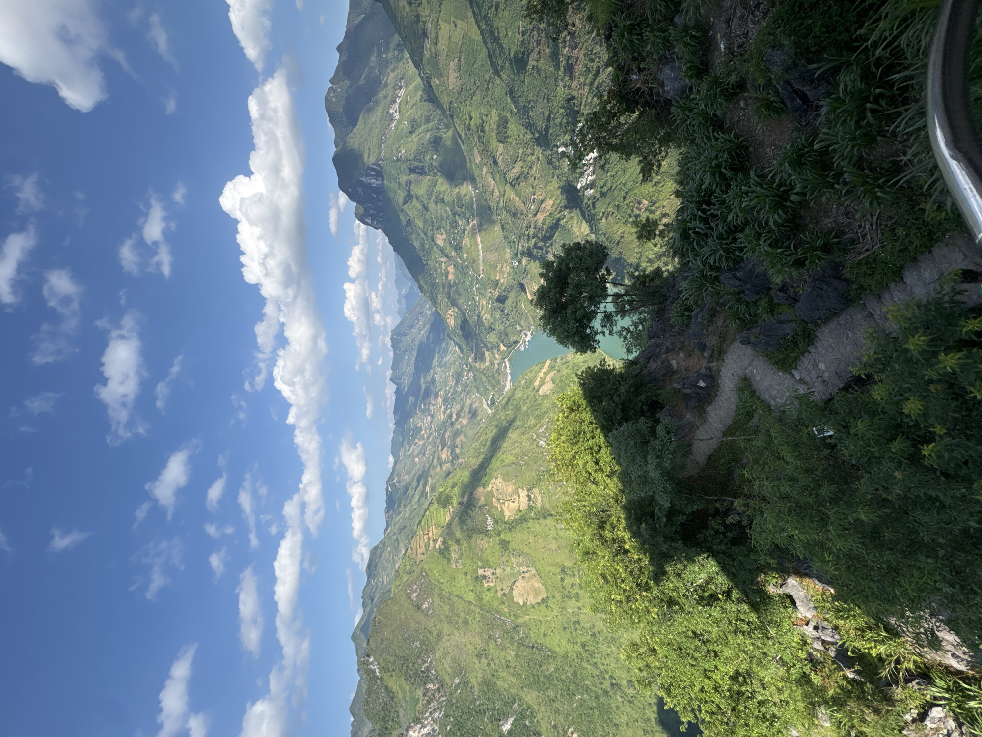 The photograph presents a view of a landscape in Đồng Văn Commune, Vietnam. The frame is dominated by steep, green, mountainous terrain. A river or lake can be seen between the mountains. The sky is visible in the left portion of the frame, filled with white clouds against a blue backdrop. The right-most edge of the image shows a portion of a stone path or wall overgrown with vegetation. A metal handrail is also visible.