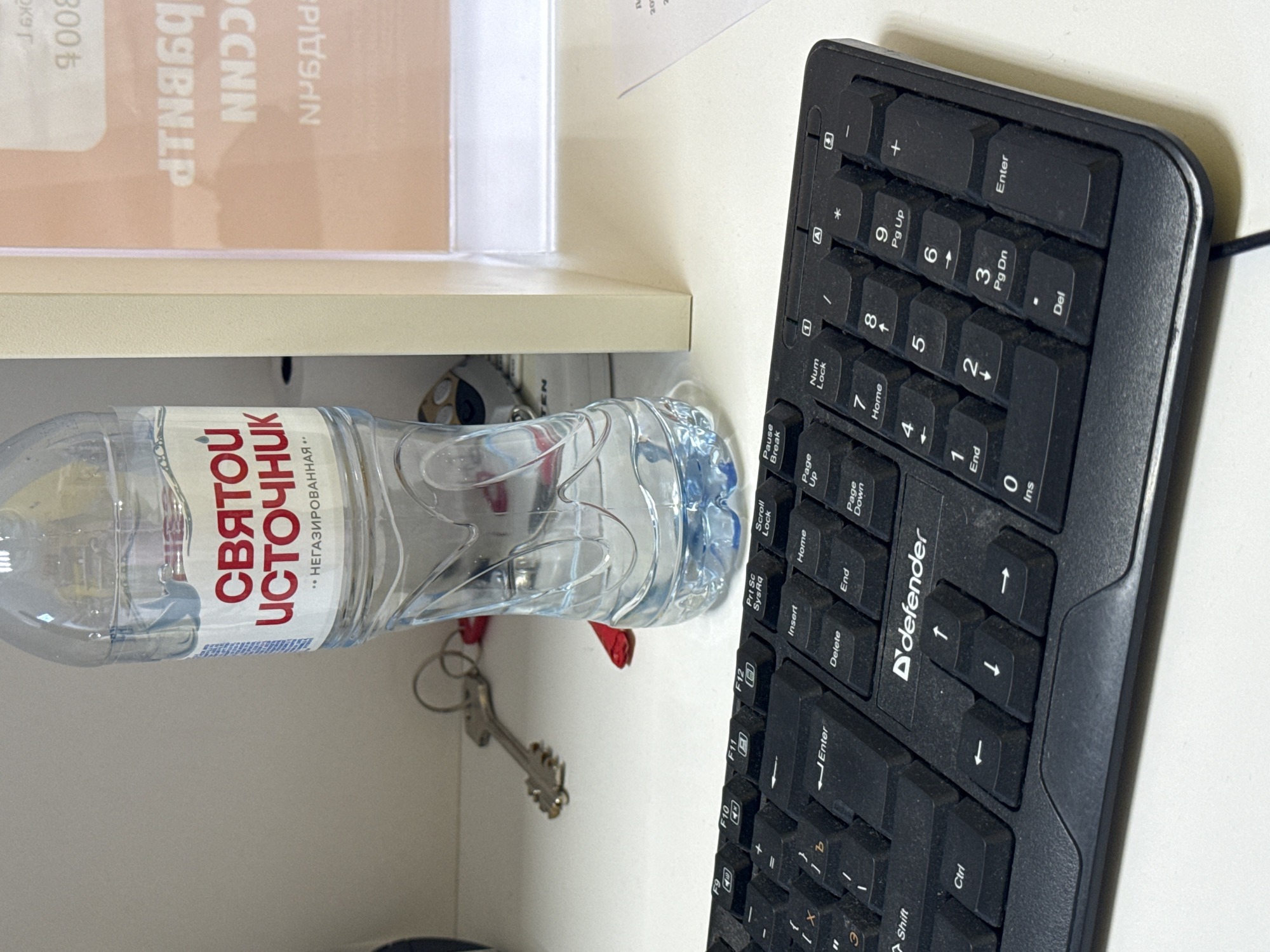 This is a top-down shot of a desk surface. Located on the desk is a water bottle with a label that reads "СВЯТОЙ ИСТОЧНИК ... НЕГАЗИРОВАННАЯ", as well as a keyboard labeled "defender." There is also a set of keys next to the water bottle. The scene appears to be a workspace. The location is unknown.