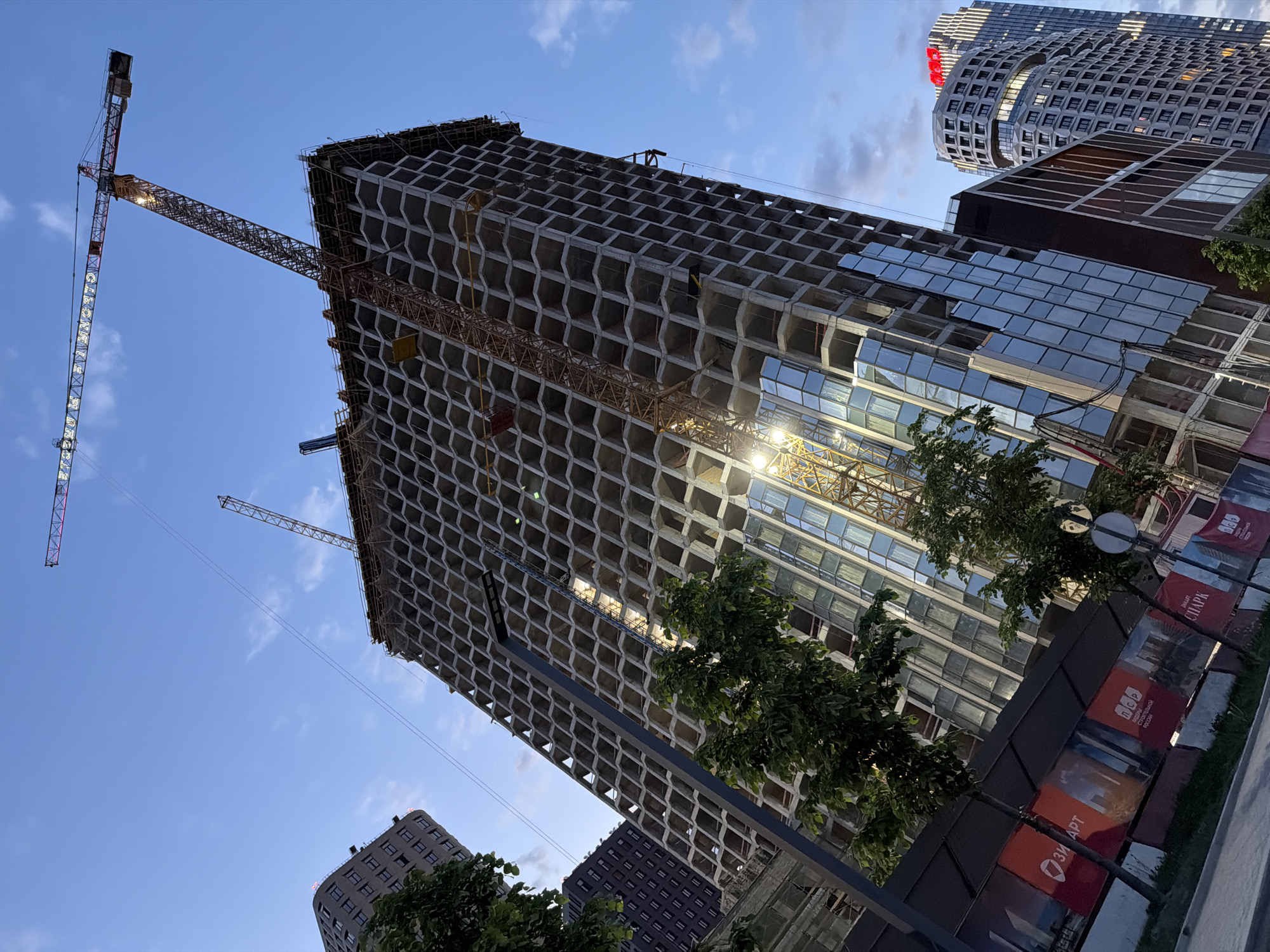 The image shows a construction site in Moscow, Russia, featuring a tall, unfinished building. The building's facade is a grid-like structure with some glass paneling installed. Two construction cranes are visible near the building. There are other buildings in the background. In the foreground, there are trees and a fence with promotional banners. The sky is blue with some clouds. The overall scene depicts urban development and ongoing construction activity in the city.