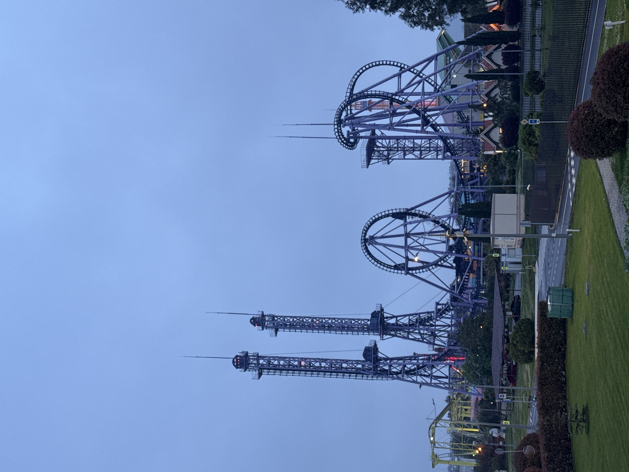 The image shows a roller coaster and drop tower amusement park attraction. The structures are constructed of steel and painted in shades of blue and purple. There is a green lawn to the right of the attractions and a road running along the right side. The sky is blue with some scattered clouds. The image appears to be taken in the evening, as it is twilight. Based on the information provided, this amusement park is located in the city of Sirius, Russia. There are no people visible.