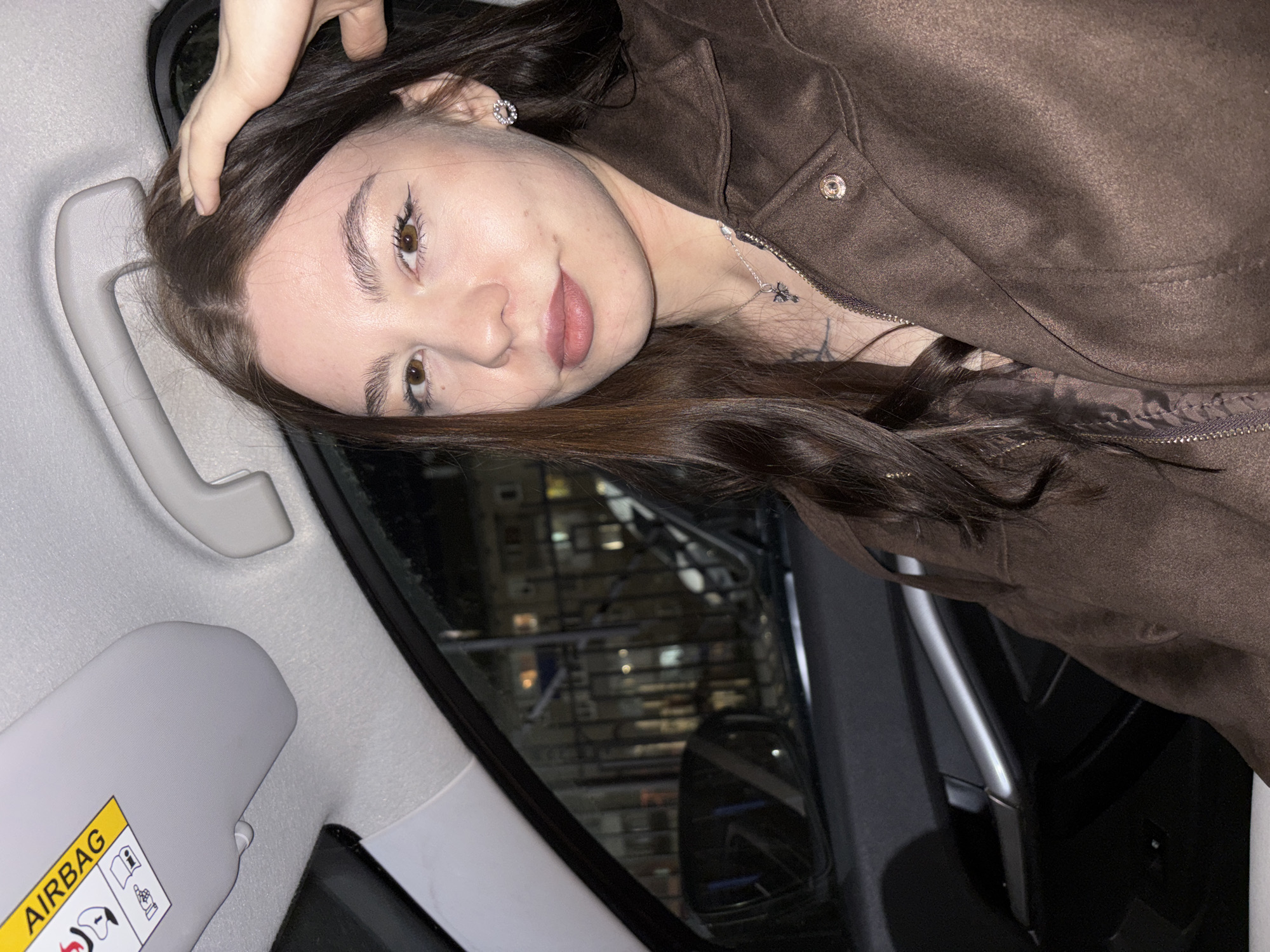 A woman with long brown hair, fair skin, and brown eyes, wearing a brown jacket and silver necklace, is seated in the passenger seat of a car, touching her hair with her left hand. The interior of the car is visible, including the sun visor with an "AIRBAG" warning label, the grab handle above the window, and part of the door. The car window shows a blurred reflection of a building at night, possibly in a city like Perm, Russia, given the building style and ambient lighting. The reflection suggests an urban environment with lighted windows.