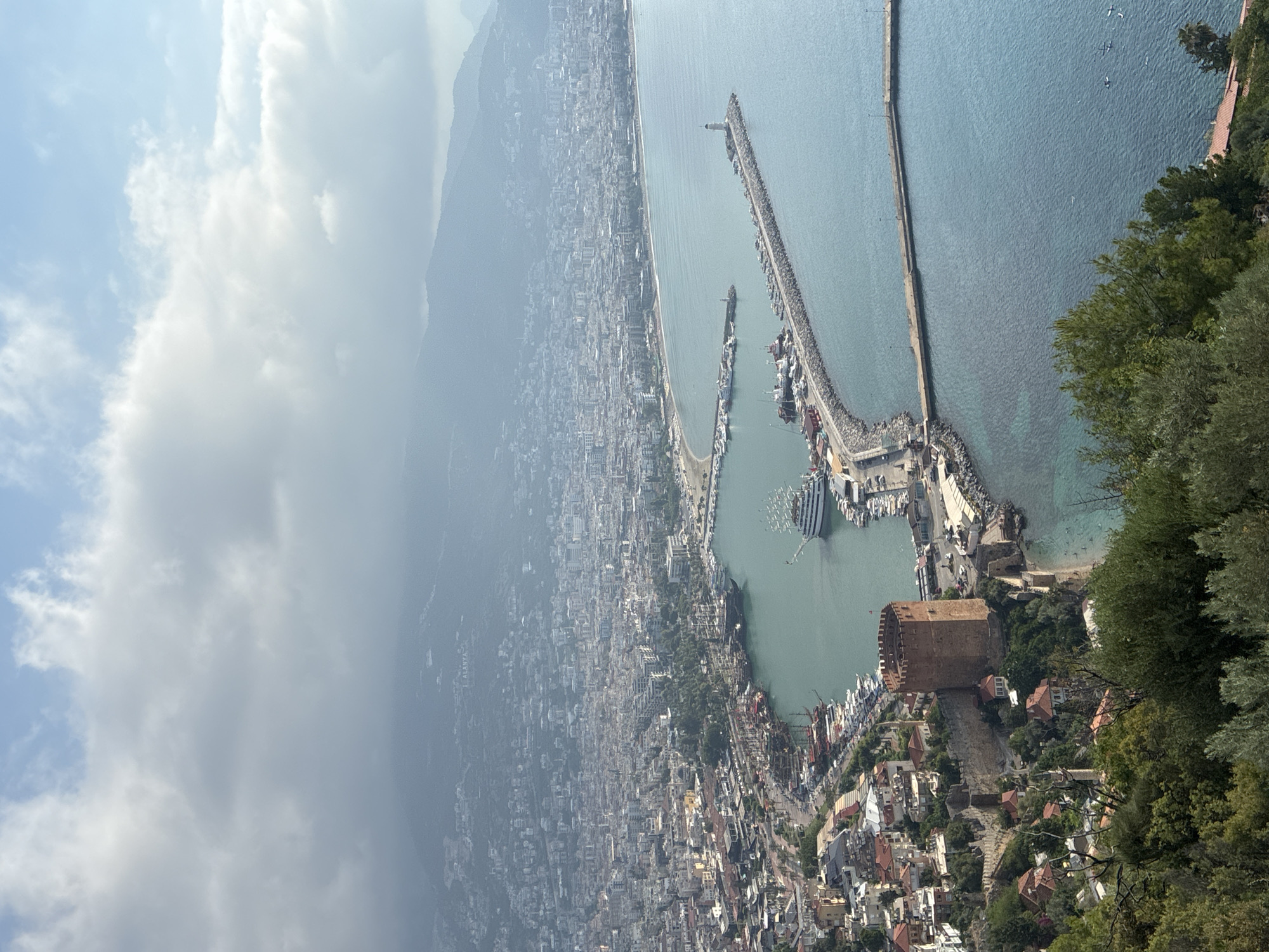 This is a high-angle, wide shot of the coastal city of Alanya, Turkey, under a cloudy sky. In the foreground, lush green trees and foliage obscure the bottom of the image. The middle ground features a prominent red-brick tower, likely Kizil Kule (Red Tower), perched on a rocky outcrop overlooking the sea. Adjacent to the tower are stone walls and a few red-roofed buildings. The harbor area is filled with numerous boats, including several large, multi-masted vessels resembling pirate ships and smaller modern yachts. A long, stone breakwater extends into the turquoise sea, with a white lighthouse situated at its end. The breakwater is lined with many small boats. Beyond the harbor, the city of Alanya stretches along the coastline, densely packed with buildings of varying sizes and colors, nestled against a backdrop of hazy, mountainous terrain. The sea is a deep blue-green, with some boats visible further out. The overall scene depicts a historic coastal city with a bustling harbor and a scenic mountain backdrop.