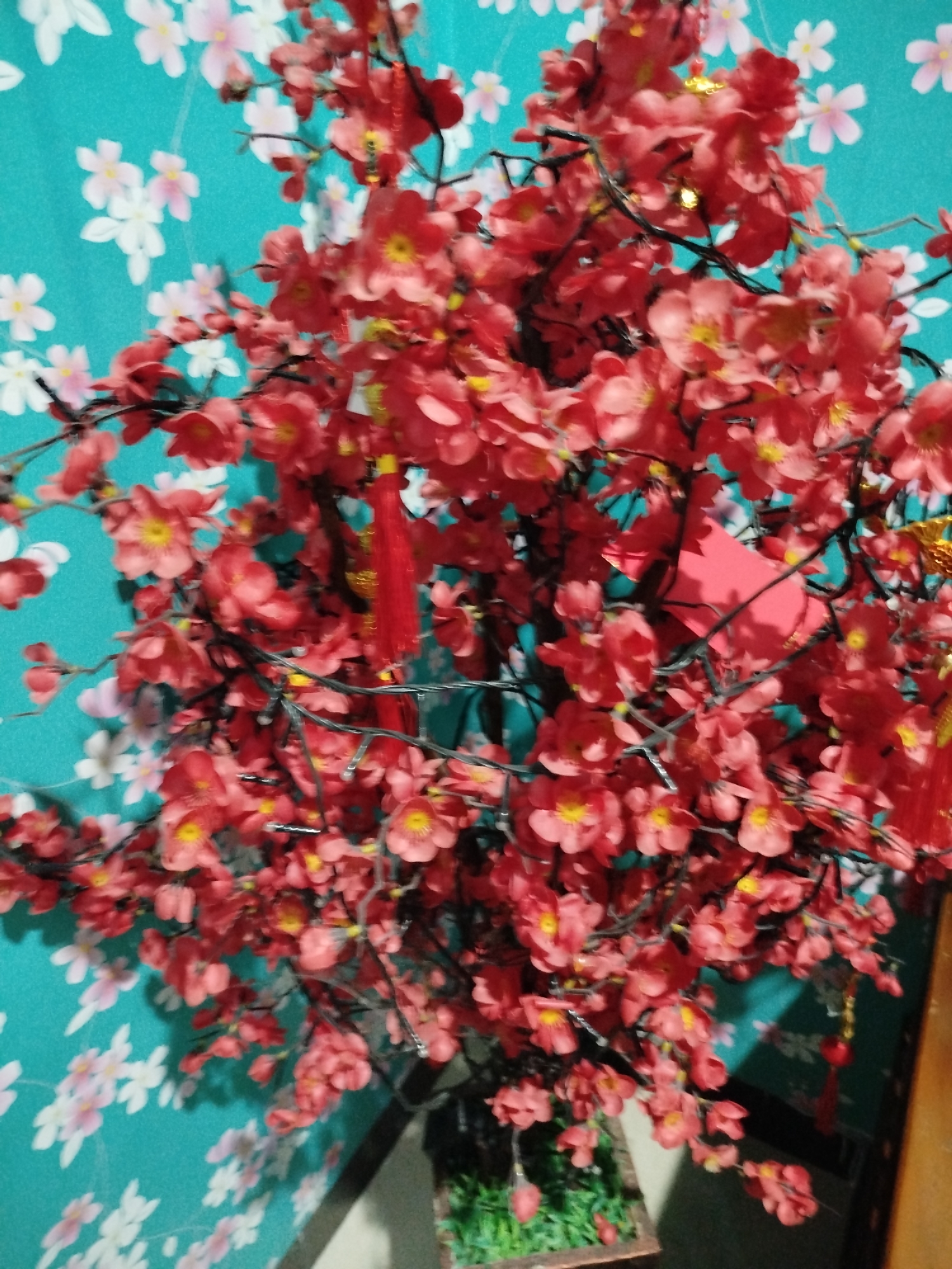 The image shows a close-up of a decorative artificial plum blossom tree with red flowers and some golden accents. The tree is adorned with a few red tassels and a small red paper decoration, possibly for a festive occasion. It is also strung with small, clear LED lights. The tree is set against a teal-colored background with a pattern of white and light pink cherry blossoms. Part of a woven planter and some artificial green grass are visible at the bottom of the frame. A wooden surface is partially visible on the right side of the image. The scene appears to be indoors, likely in a residential setting in Bandungrejo, Indonesia, given the context provided. There are no people or animals present in the image.