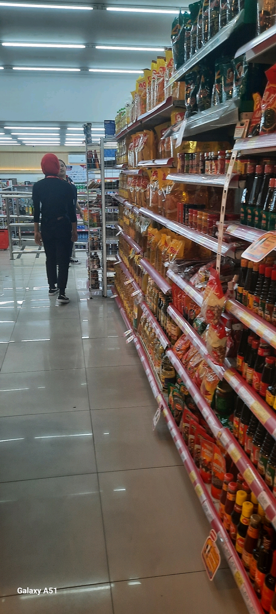 The image depicts an interior scene of a grocery store aisle in Wlingi, Indonesia. A person wearing a red hijab and a black outfit is walking away from the camera, down the aisle. The aisle is lined with shelves filled with various packaged food items, predominantly sauces and cooking oil, in pouches and bottles. The flooring appears to be tiled and well-lit by overhead fluorescent lights. In the background, other shelves and products are visible, along with what appear to be metal shopping carts or display fixtures. The overall environment suggests a typical retail setting.