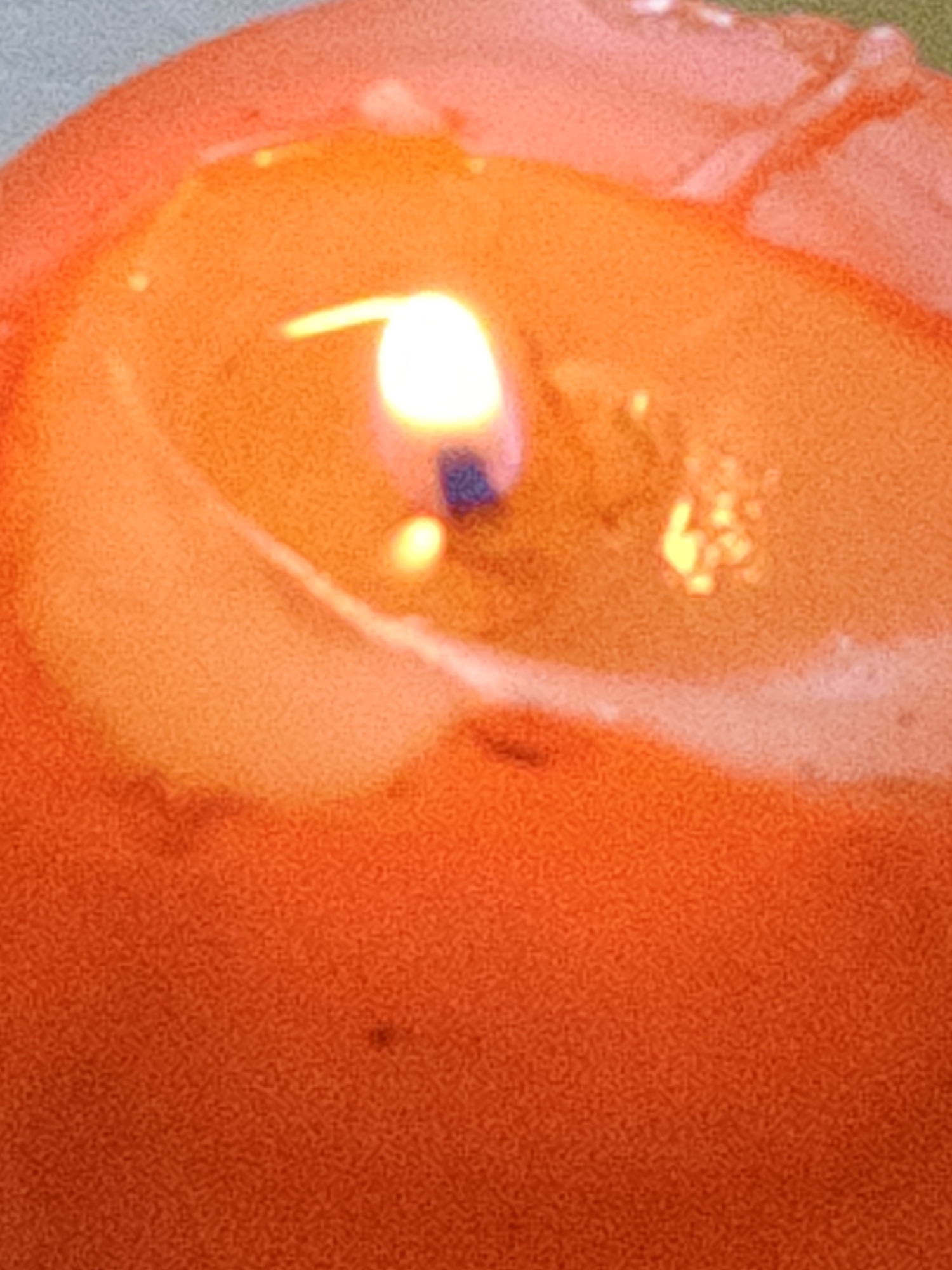A close-up, slightly blurry image captures a lit orange candle. The flame is bright white at its core, transitioning to yellow and then orange at the edges, casting a warm glow. The melted wax forms a wavy surface around the wick, with some lighter patches suggesting reflections or variations in the wax. A faint, dark speck is visible on the wax surface. The background is out of focus, appearing as a textured, light gray or pale green area at the top left. The overall impression is one of warmth and gentle illumination.