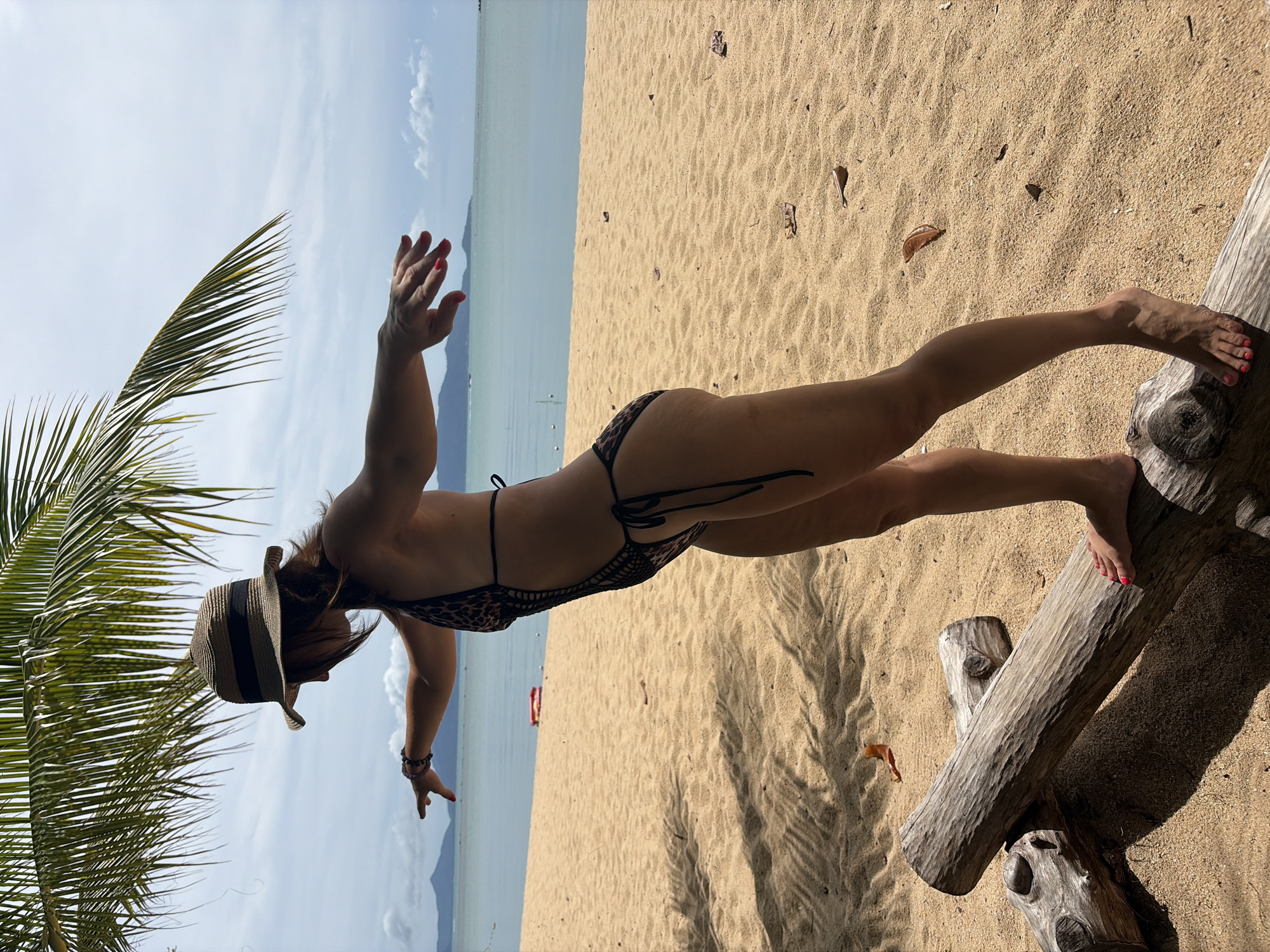 A woman wearing a leopard print swimsuit and a straw hat stands on a beach in Baan Bo Phut, Thailand. She is balancing on a log, with her arms extended. The background shows the ocean, a cloudy sky, and palm fronds. The sand is golden and scattered with dry leaves. The woman has dark hair and is facing to the left of the frame.