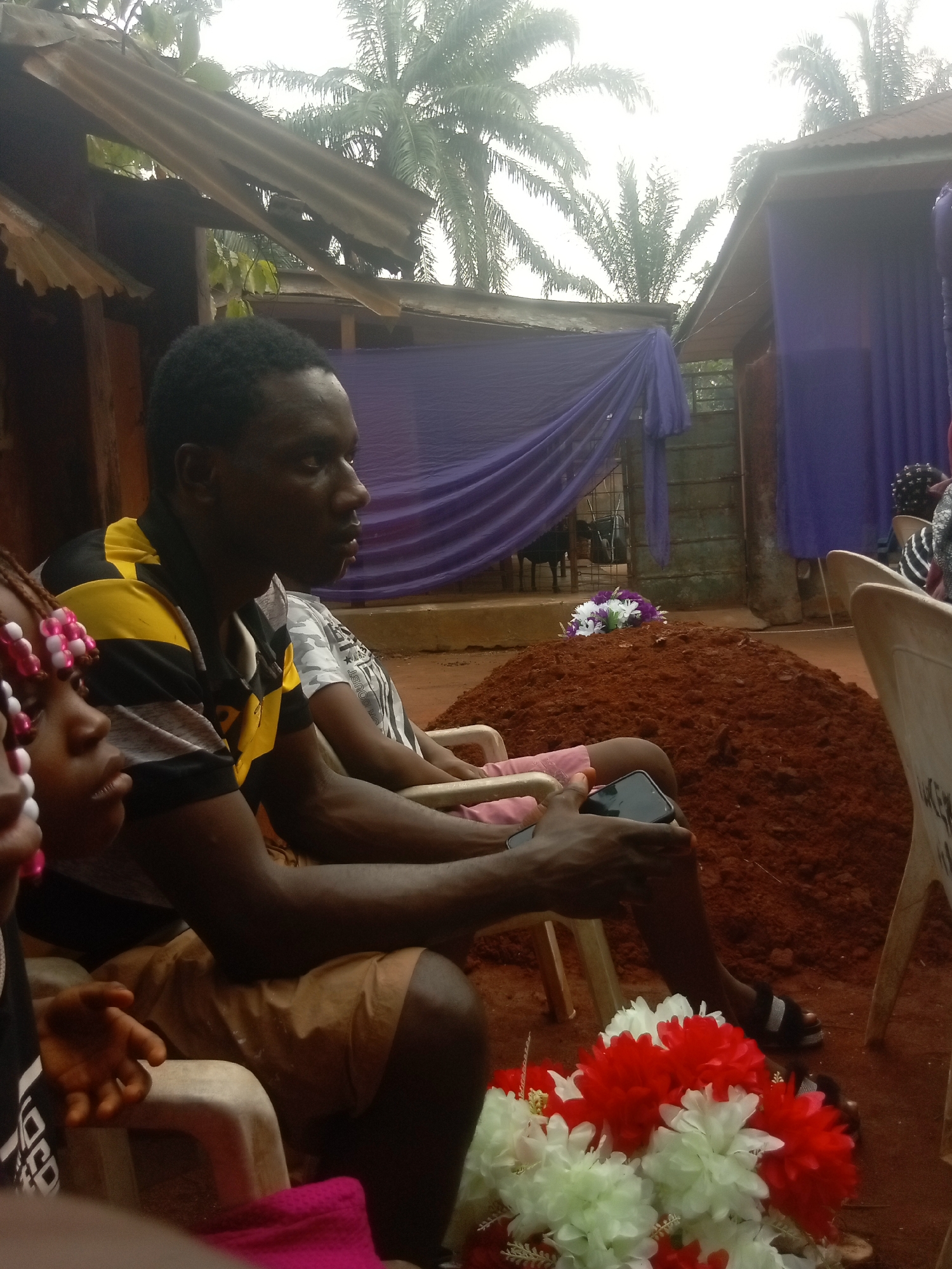 The image shows an outdoor scene in what appears to be Igbo-Ukwu, Nigeria. A freshly dug grave mound of reddish-brown soil is centrally positioned. In the foreground, a man is sitting on a plastic chair, holding a phone. A small girl with beaded braids sits partially visible beside him. Additional people are seated in chairs behind them. Artificial flowers adorn the ground near the chairs and are on the grave. The background includes buildings with corrugated metal roofs and draped purple fabric. Palm trees are visible in the sky above the buildings.