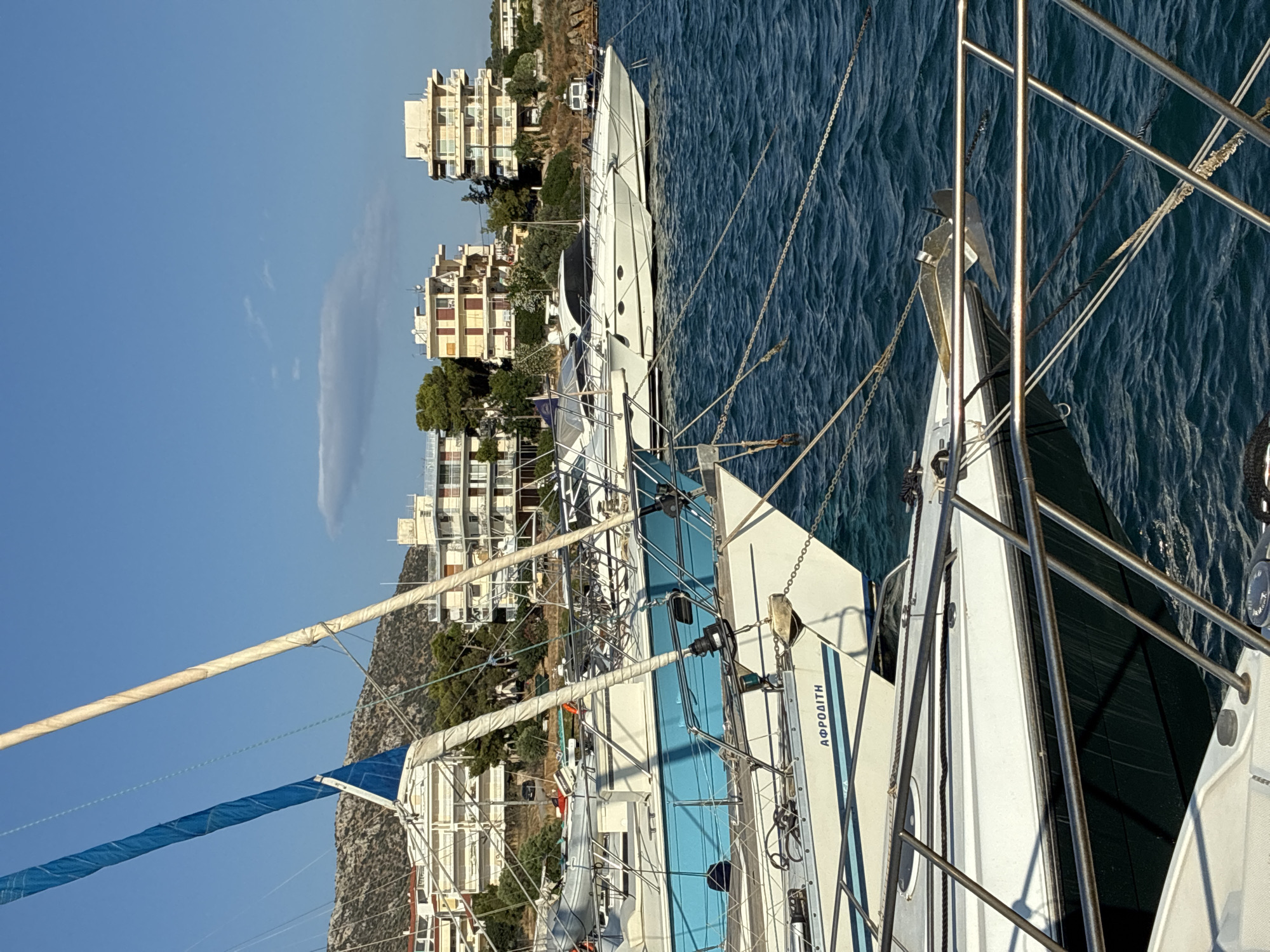 The image shows a harbor in Limenas Markopoulou, Greece, with several boats moored in the blue water. In the foreground, a white sailboat with a blue stripe on its hull is visible, with the name "ΑΦΡΟΔΙΤΗ" (Aphrodite) written on its side. Another sailboat with a blue sail is partially visible behind it. In the background, there are several white buildings with balconies and red-tiled roofs, set against a backdrop of a rocky hill and a clear blue sky with some scattered clouds. The water is choppy, indicating a slight breeze. The scene appears peaceful and serene, characteristic of a Mediterranean coastal town.