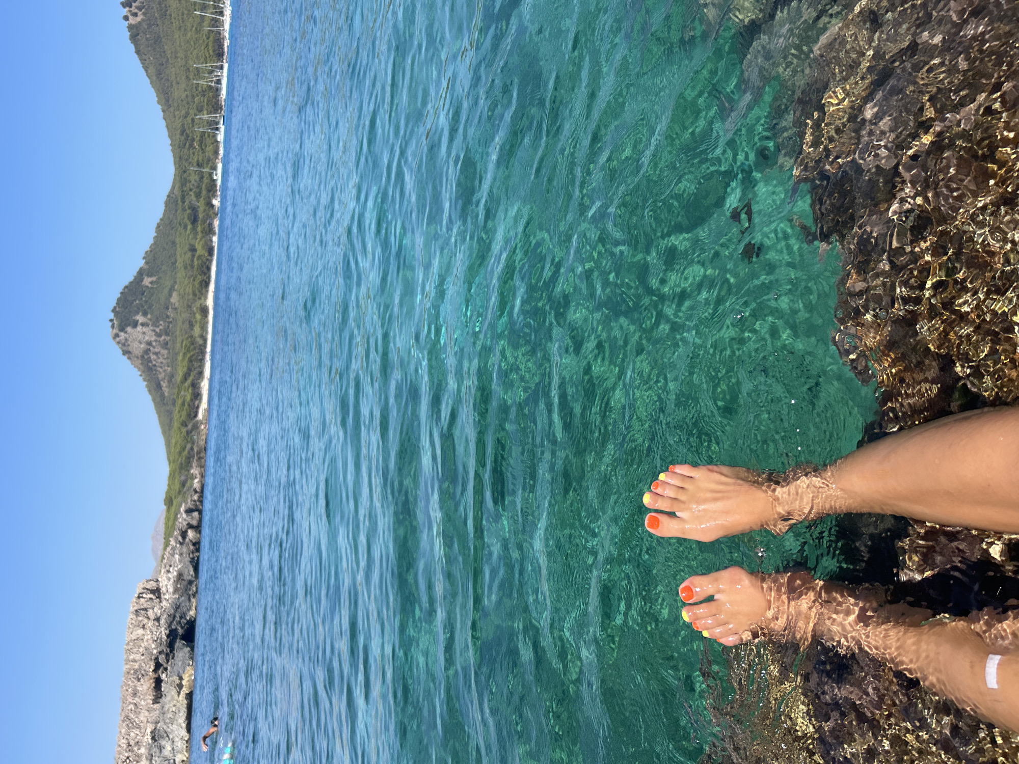 In Cala Lliteres, Spain, a person's bare feet, with orange and yellow painted toenails, are submerged in clear turquoise water. The feet rest on dark, rocky surfaces partially submerged at the edge of the water. The water is clear enough to reveal the texture of the rocks below the surface. In the background, the bright blue sky meets the horizon where green, tree-covered hills rise, with a bay dotted with sailboats visible in the distance. The scene conveys a sense of relaxation and a beautiful coastal environment.
