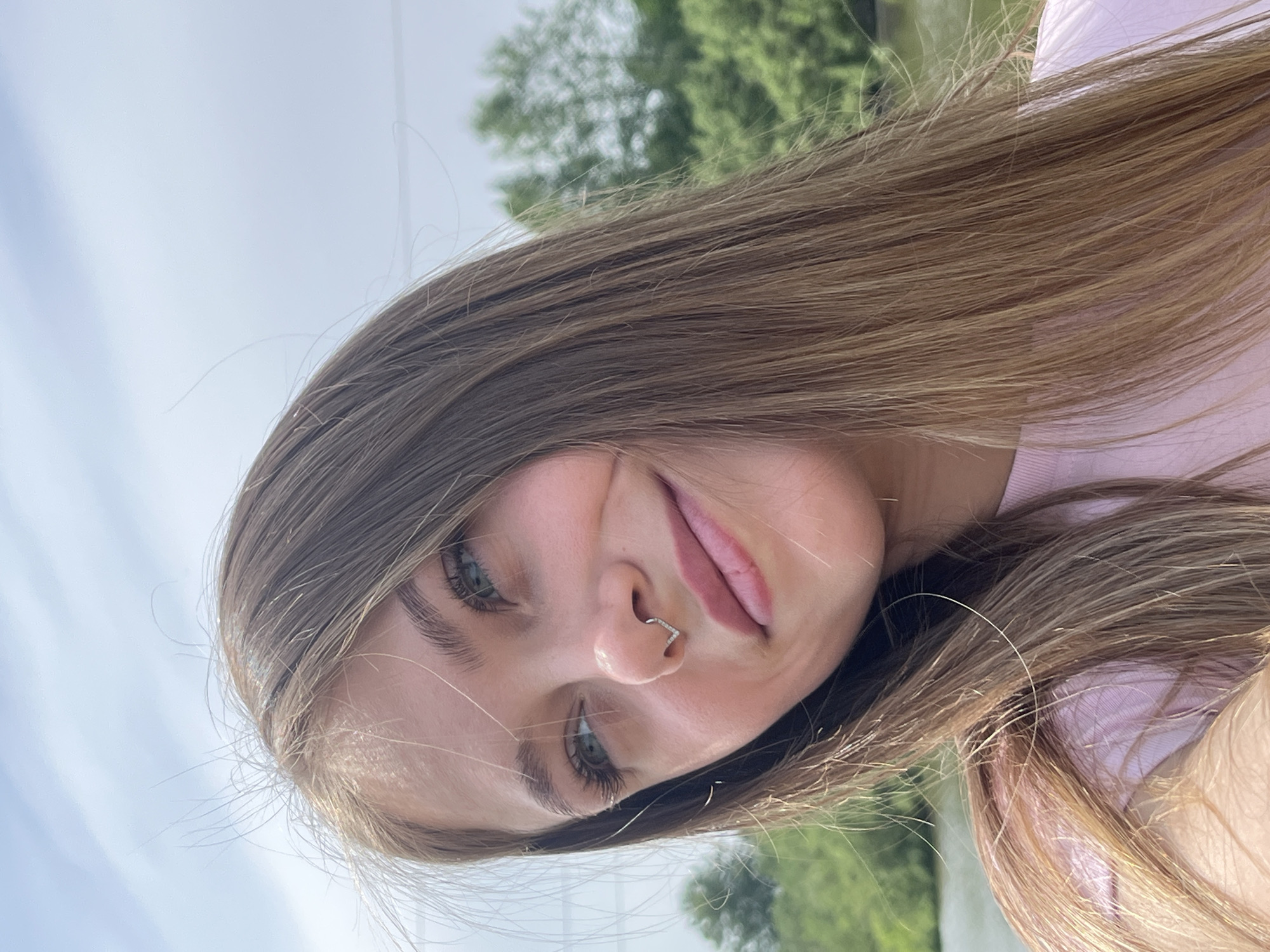 A young woman with light brown hair and a nose piercing is photographed from a slightly low angle. She is wearing a light pink top. The background consists of a light blue sky and green trees, suggesting an outdoor location. No specific location within Фоки, Russia can be discerned.