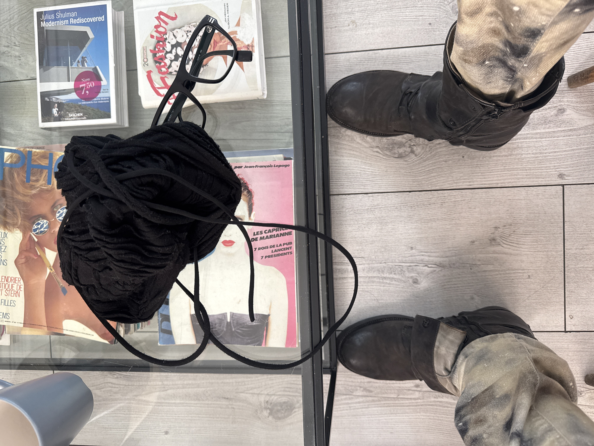 An overhead shot captures a pair of feet in dark, worn boots and light-colored, possibly distressed, pants, positioned on a light wooden floor. The feet are seen next to a glass-top table or display case.
On the table, various items are arranged. A large, textured bundle of black yarn or fabric is prominently placed, partially obscuring a magazine cover featuring a woman wearing artistic sunglasses. Below this, a light blue mug is partly visible. To the right, another magazine cover with a pink background depicts a woman in a black bustier.
Further back on the table, a book titled "Julius Shulman Modernism Rediscovered" by TASCHEN is visible, featuring a modernist building on its cover and a "Now 7,50" price sticker. Black-rimmed eyeglasses rest on a stack of books, one of which has "Fion" and names "JIM HEIDMANN" and "ALISON A. NIEDER" visible. The eyeglasses have a partially visible "BURBERRY" inscription.
Visible text fragments from magazines include "PHOTOGRAPHIE", "par Jean-François Lepage", "LES CAPRICES DE MARIANNE", "7 ROIS DE LA PUB LANCENT", and "7 PRESIDENTS," suggesting French content and aligning with the Paris, France context. The scene depicts an indoor setting, likely a home or creative space, with no specific activity beyond the arrangement of these personal and cultural items.