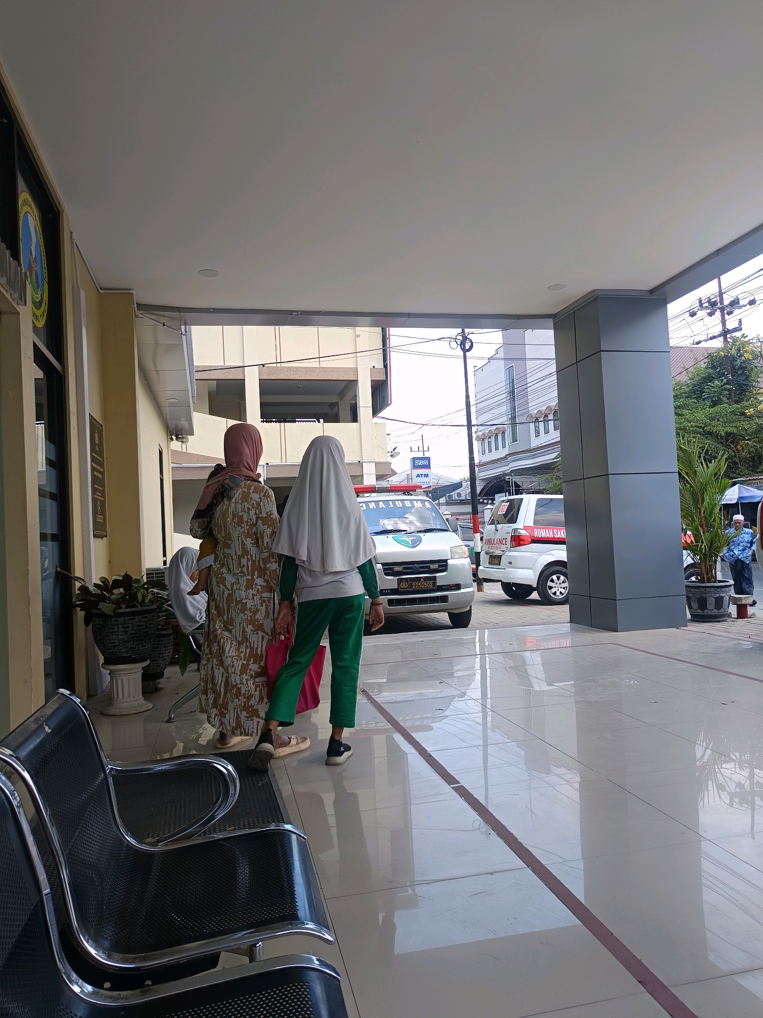 The image depicts the exterior waiting area of a hospital or medical facility in Bondowoso, Indonesia. In the foreground, black metal benches with chrome accents are visible. A woman wearing a pink hijab and a patterned dress, carrying a child, stands in the middle ground. To her right, another woman, wearing a white hijab and green track pants, holds a pink bag. Behind them, two ambulances are parked. One is white with red and black lettering that reads "RUMAH SAKIT" (Hospital) and "AMBULANCE". The other ambulance, in front, is also white and has a logo on its side. The building's facade is yellow with a covered walkway. The ground is tiled with a reflective surface. Electrical poles and wires are visible in the background, along with other buildings and trees, suggesting an urban environment.