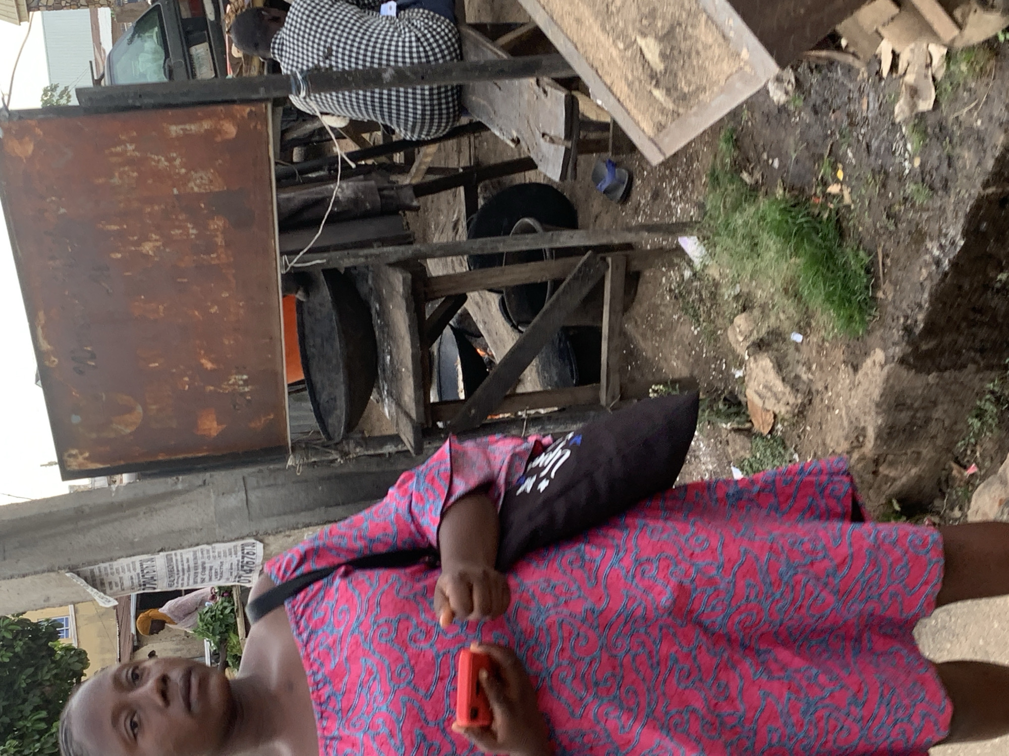 This media file captures an outdoor scene, likely a street or market area in Jos, Nigeria, characterized by makeshift structures and daily activity. The image is taken from a slightly elevated perspective, looking downwards, and is rotated, depicting subjects mostly on their sides.
In the foreground, a woman stands prominently, wearing a vibrant pink dress with a blue swirling pattern. She carries a black shoulder bag that has some white text on it, partially obscured but appearing to read "I'M A W..." or "I'M A Y...". In her right hand, she holds a small, rectangular red object, possibly a mobile phone or another device. Her expression is neutral, and her gaze is directed slightly upwards and towards the left.
The background reveals a complex arrangement of objects and people. Behind the woman, there's a rusty, rectangular metal sheet or sign, possibly part of a stall or structure. Further back, another individual is seated behind a rough wooden framework, wearing a black and white checkered shirt. The ground around them is uneven, appearing muddy in places, with sparse patches of green grass and some discarded items, including what looks like a blue flip-flop. Visible within the wooden structure are black objects, possibly tires or barrels.
In the mid-ground, a concrete wall or pillar is adorned with a white sheet of paper containing visible black text, including phone numbers "070476767" and "0704767670". Below this, another person with a yellow head covering is partially visible, further suggesting a bustling, communal environment. The lighting appears soft, indicating either an overcast day or perhaps early morning/late afternoon, as there are no harsh shadows. The overall impression is one of everyday life in a busy, informal urban setting.
