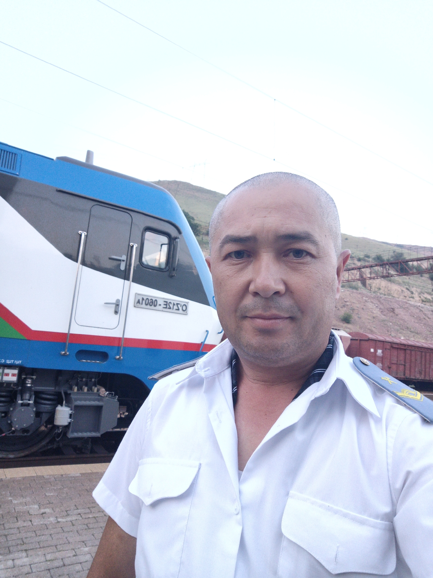 The image features a man in a white uniform standing in front of a train. He is bald with a light complexion and appears to be middle-aged. He is wearing a white collared shirt with epaulets on his shoulders, indicating a position of authority. A dark and white striped band is visible around his neck, possibly part of his uniform or an accessory.
The train behind him is modern and sleek, with a blue, white, and red livery. The word "UZBEKISTAN" is visible in text on the train. The setting appears to be a train station or depot, with brick paving on the ground in the foreground and tracks visible. In the background, there are hills and what looks like a bridge or overpass structure. The sky is bright, suggesting it is daytime, possibly late afternoon or early evening given the lighting. The overall mood of the image is neutral, with the man looking directly at the camera. There is a small plaque on the side of the train with text that appears to be in a Cyrillic script, and the letters "O.SISE - OEOV." Another piece of text is visible on the lower part of the train.