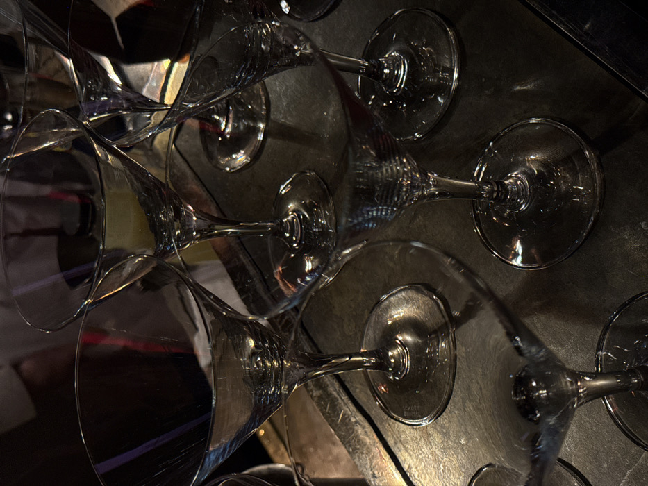 Several wine glasses are arranged haphazardly on a dark, reflective surface. The glasses vary in position and overlap, creating a complex interplay of light and shadow. The glasses appear clean and unused, suggesting they are awaiting service. The background is indistinct, emphasizing the glasses as the main subject. The scene evokes a sense of anticipation and elegance.
