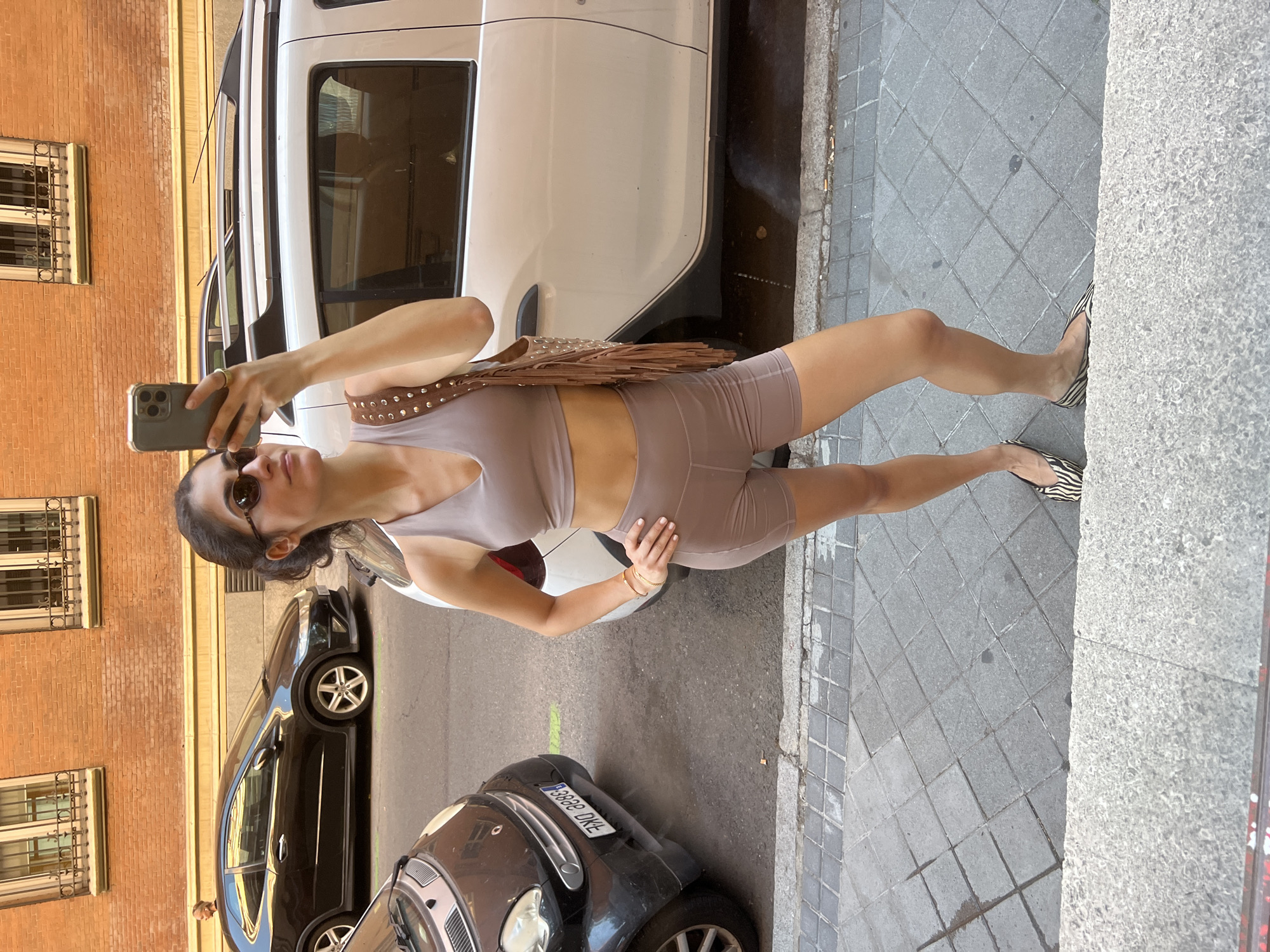An urban street scene unfolds in bright daylight, featuring a woman taking a full-body selfie. She stands with one leg slightly extended and a hand on her hip, holding a smartphone up to capture the image. She wears dark sunglasses and a matching light taupe or brown athletic two-piece set, comprising a crop top and cycling shorts. A brown handbag, adorned with silver studs and fringe, is slung over her shoulder. Her flat shoes feature a distinctive black and white zebra print.
The background reveals a bustling street environment. Immediately behind her is a white van or SUV, reflecting the surroundings. Further down the street, a black hatchback car is parked. In the foreground, a small, dark-colored car is visible, its license plate "3868 DKL" clearly readable. The woman is on a gray paved sidewalk made of square tiles, bordering a textured concrete curb and the asphalt street. To her left, a building with warm reddish-brown brickwork and windows fitted with decorative wrought iron bars lines the street. The bright sunlight and architectural details are consistent with a European city setting, aligning with Madrid. The photo is taken from a slightly elevated and tilted perspective.