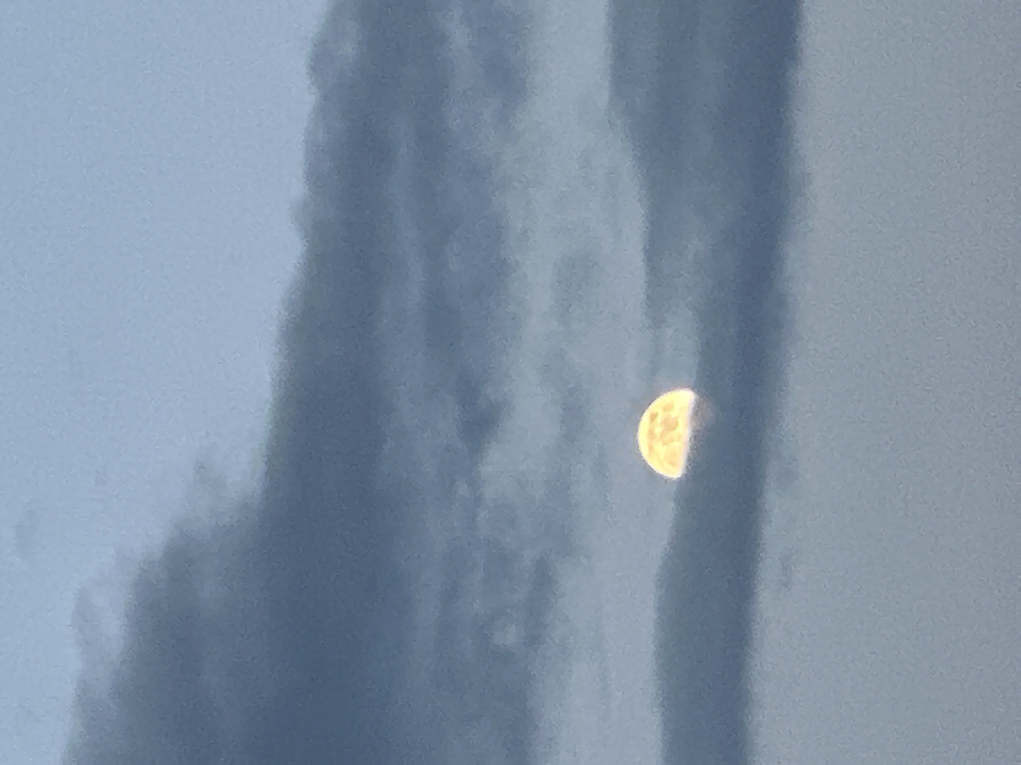 The image depicts a half-moon partially obscured by dark clouds. The moon is on the right side of the image and appears to be illuminated, with visible craters and texture. The clouds are gray and appear to be dense, with varying shades suggesting depth and movement. The background is a light blue, possibly indicating the sky. There are no people, subjects, interactions, or additional objects visible in the scene. The location is not provided.