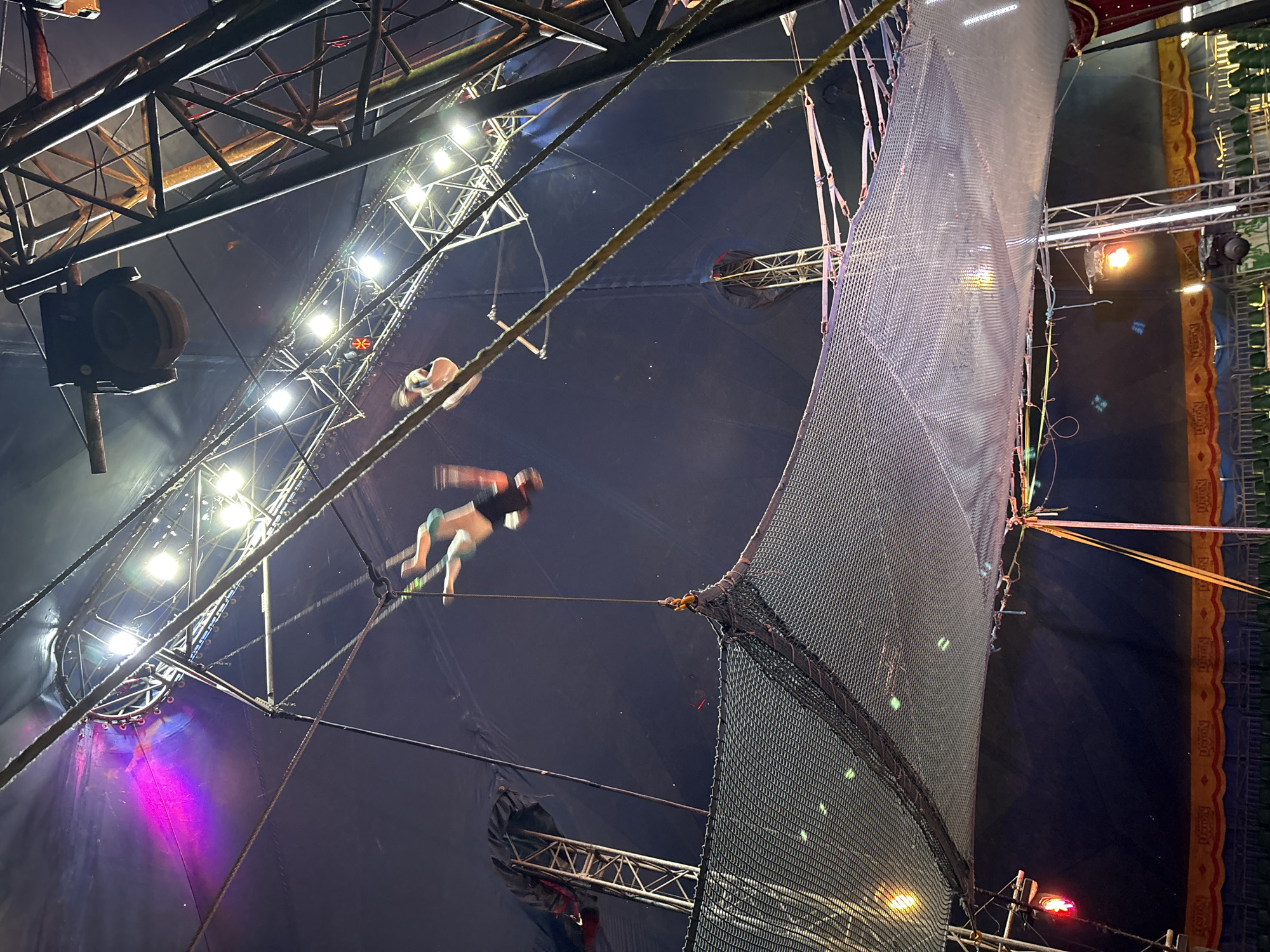 The image shows a scene inside a circus tent in Tejalpa, Mexico. The view is directed upwards towards the tent's ceiling. Visible are acrobats performing on trapezes high above the ground. A safety net is also present, running along the perimeter of the performance area. There are lights illuminating the space, and the rigging and structural elements of the tent are visible. The tent is a dark color.