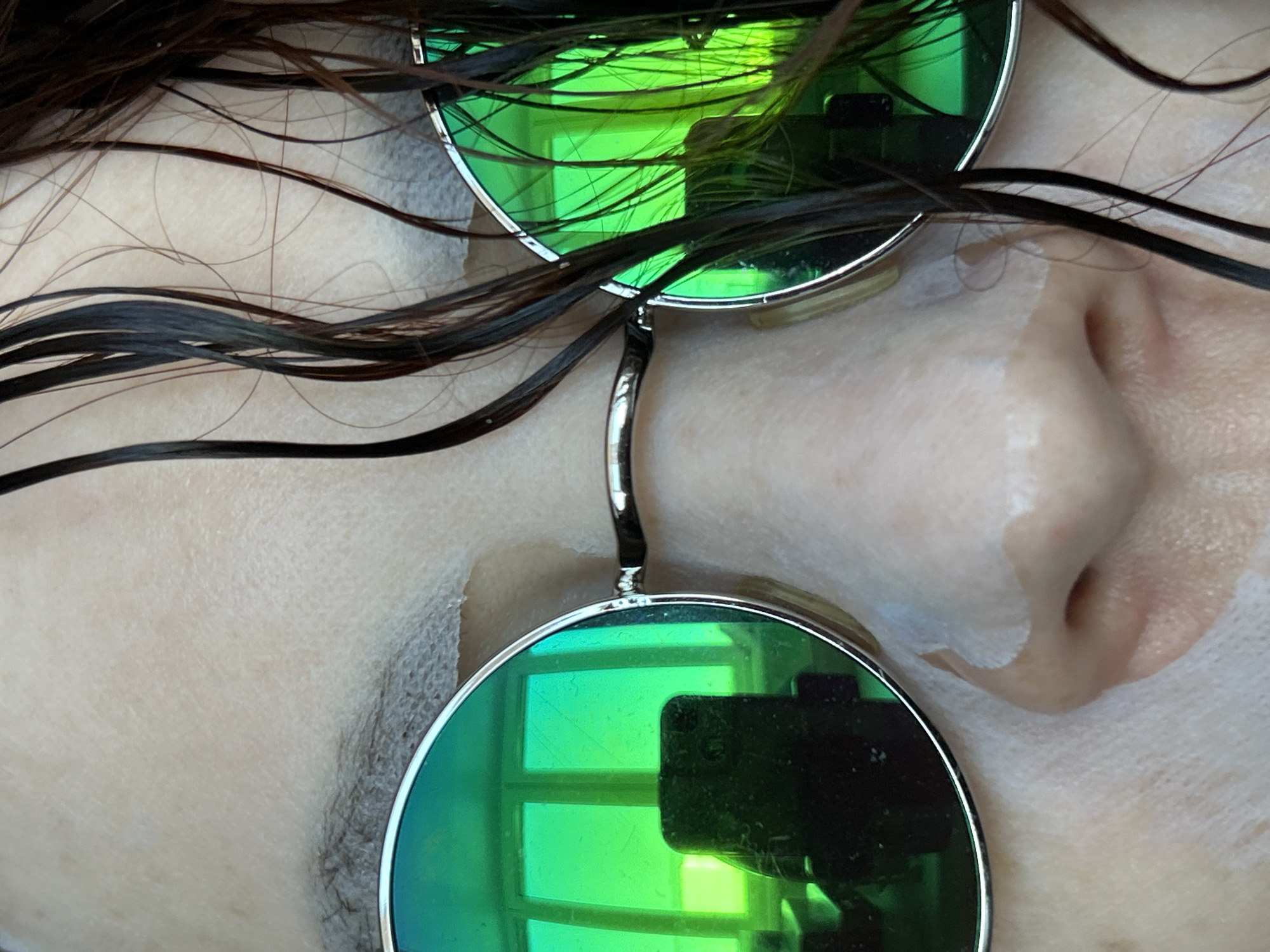 A close-up shows a person's face covered with a white sheet mask and wearing round, green-tinted sunglasses. Wet, dark hair is draped across the face. The sunglasses reflect a window and a dark object, possibly a phone or camera. The person's nose is visible beneath the mask, and part of an eyebrow is visible above the glasses. The location is unknown, the question about Chaikovsky is irrelevant.