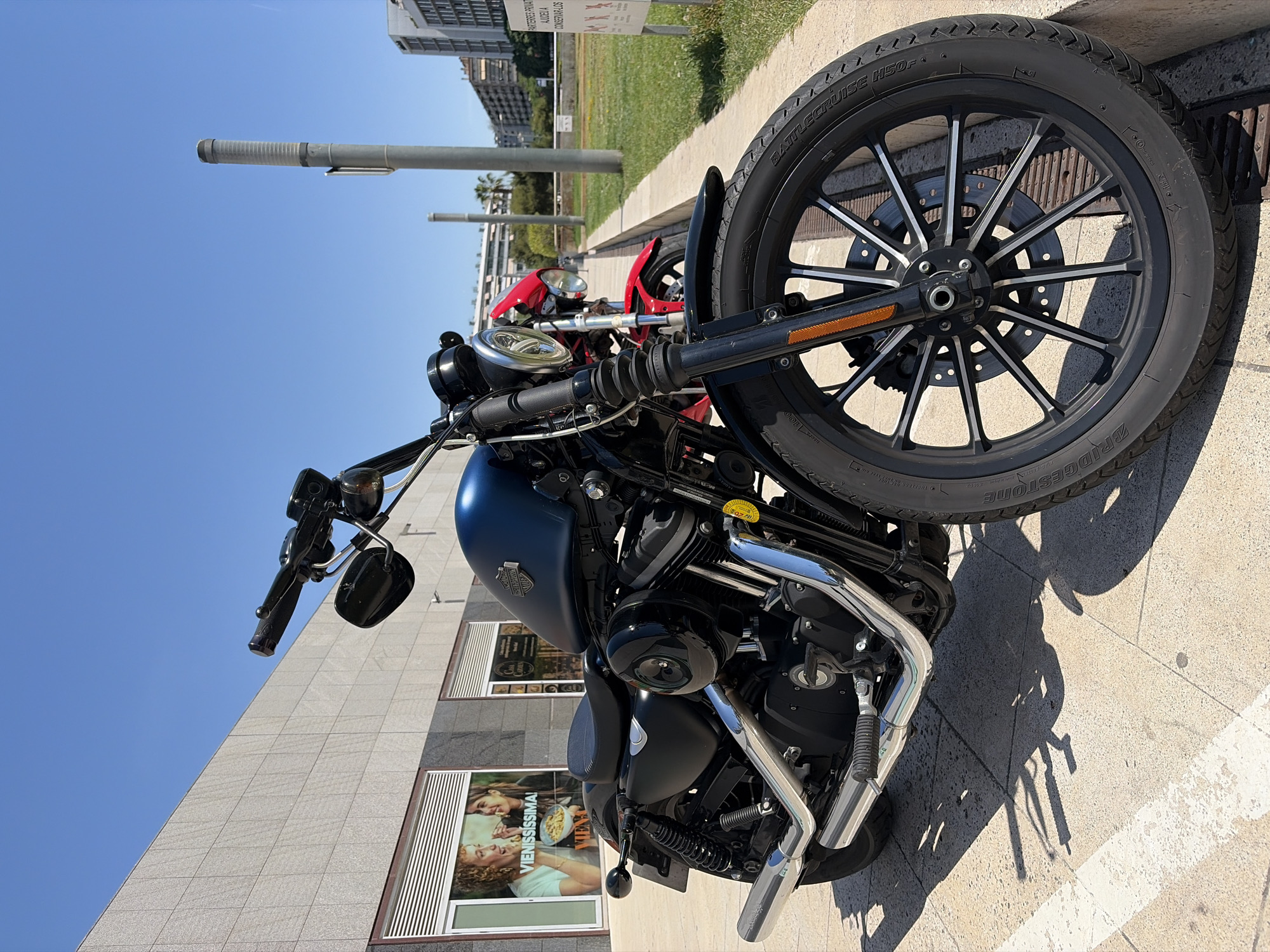 The image shows a black and chrome Harley-Davidson motorcycle parked on a sidewalk. Another red motorcycle is partially visible behind it. The background includes a building with advertisements in the windows and a grassy area with street lights. The location appears to be in L'Hospitalet de Llobregat, Spain, based on signage visible in the background. The lighting suggests a sunny day.