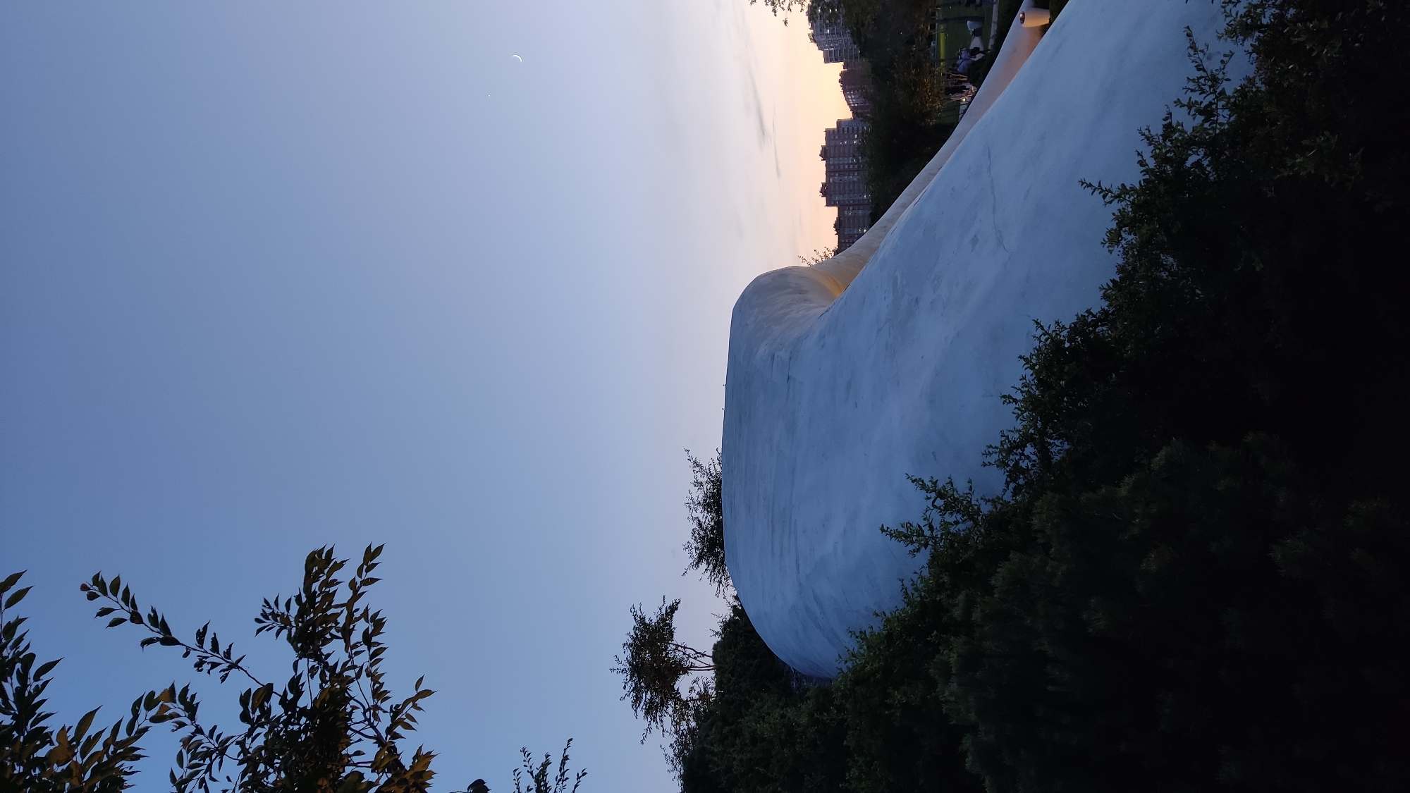 The image depicts an outdoor scene at dusk, with a clear sky gradually transitioning from a pale blue at the top to warmer hues of orange and pink near the horizon. A sliver of a crescent moon is visible in the upper right quadrant of the sky. In the foreground, a large, smooth, white, curved structure dominates the lower half of the frame, partially obscured by dark green foliage and trees on the left and bottom. This structure appears to be part of an architectural design. In the background, beyond the foliage and structure, a cluster of apartment buildings is visible against the dusky sky. The scene suggests a park or public space within a city. The specific location is the "Tachka" monument, a distinctive modern sculpture resembling a stiletto heel, situated in Krasnodar, Russia.