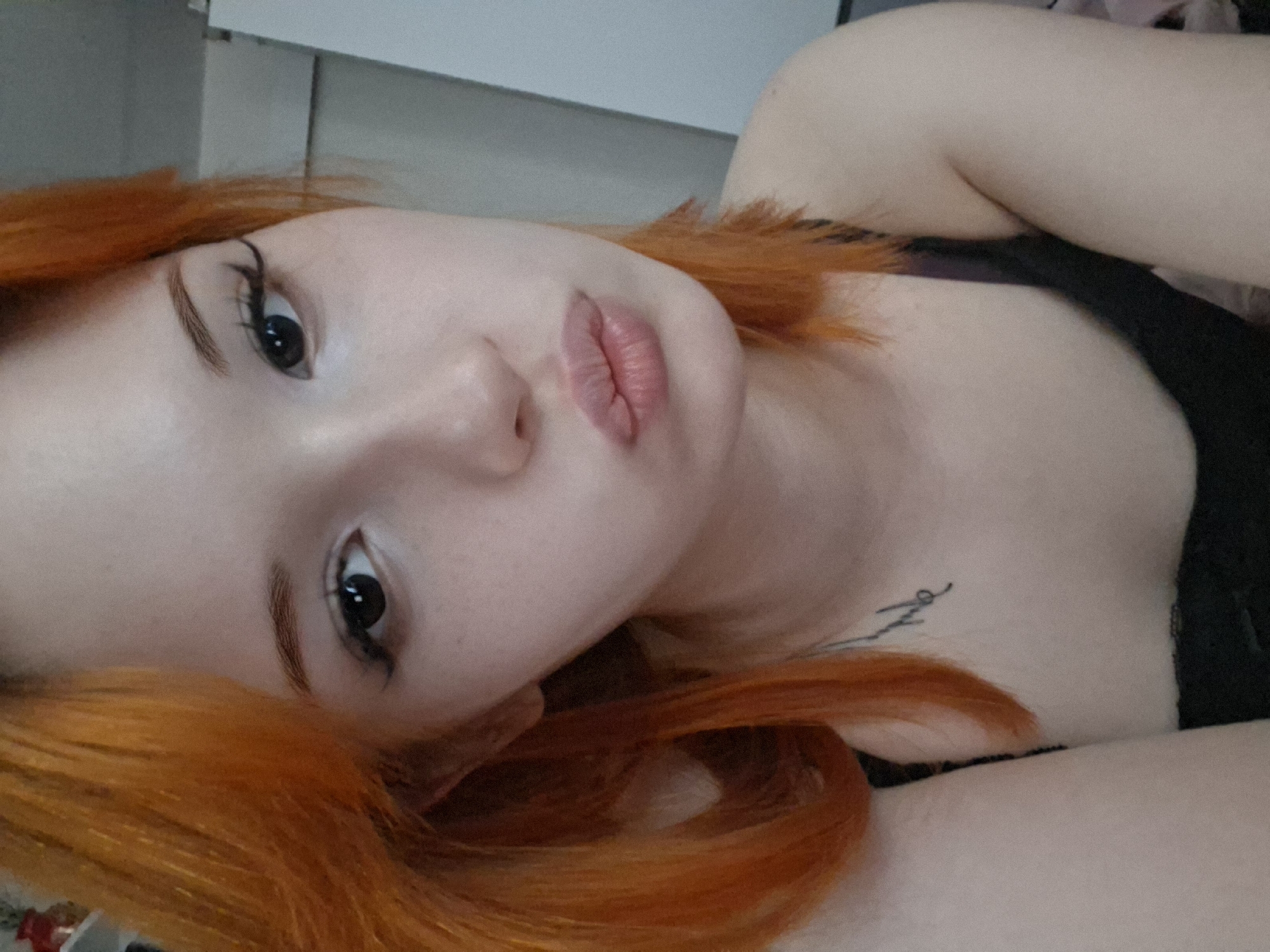 A close-up, angled, portrait of a fair-skinned woman with bright orange hair lying down. She has dark eyeliner and light pink lips. A dark garment is visible on her upper body. A tattoo is visible on her chest, though the design is not clear. The background is a plain white or light-colored wall. The location in Chaikovsky, Russia, is not discernible from the image.