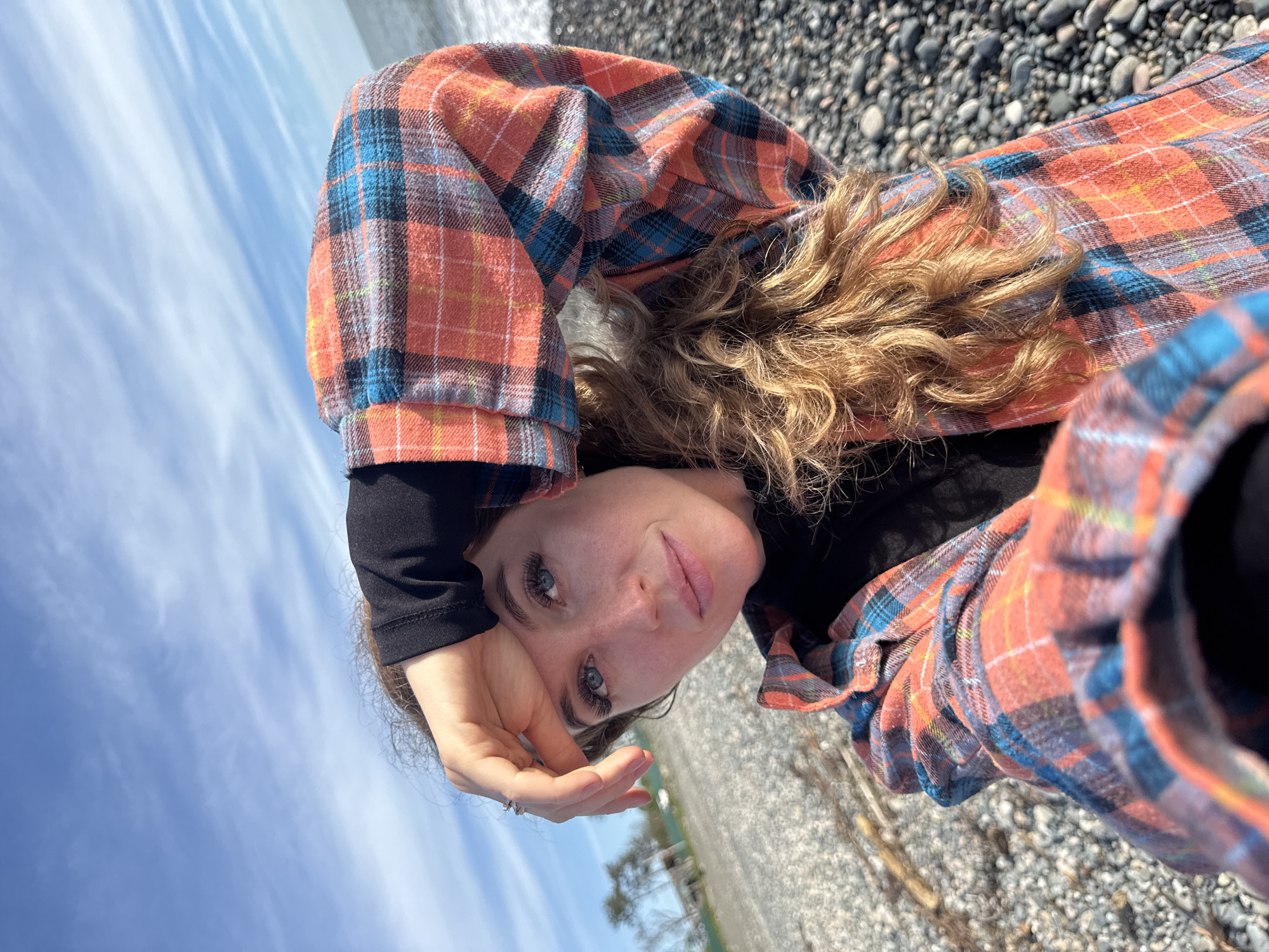 A young woman with light skin and curly blonde hair is lying on a pebble beach in Gulrypsh, Abkhazia. She is wearing a black long-sleeved shirt under an open orange and blue plaid flannel shirt. Her head is tilted, and she is looking towards the camera with a slight smile. Her right hand is raised near her face, obscuring the sky, which is partially visible in the background. The pebbles of the beach are grey and white. There is vegetation faintly visible in the background, suggesting a coastal area. The image appears to be a selfie.
