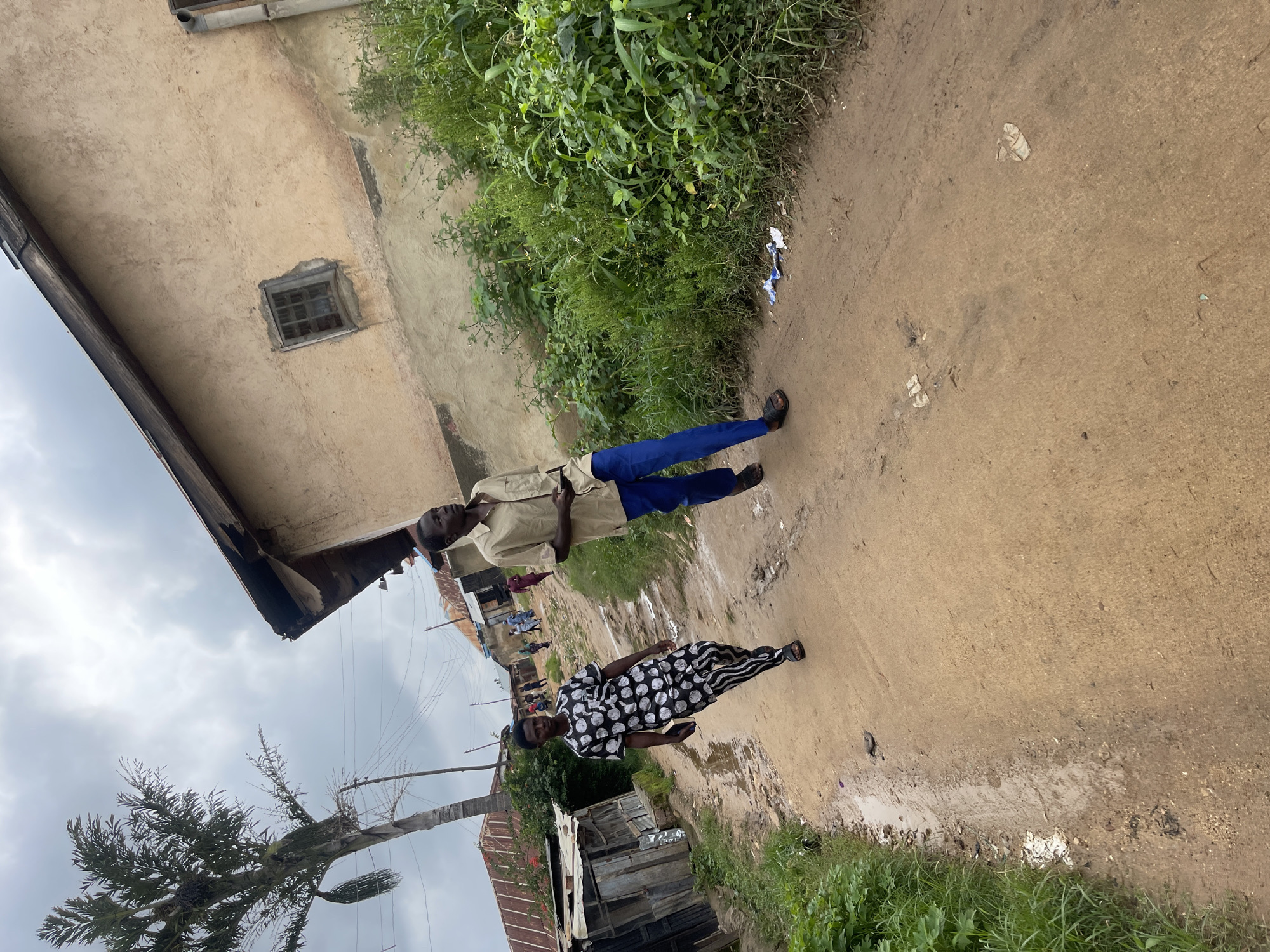 The image depicts a dirt road in an urban or semi-urban environment, likely in Jos, Nigeria, judging by the architecture and vegetation. Two men are walking on the road. The man in the foreground on the left wears a black and white patterned traditional outfit, including a short-sleeved top and trousers, and is wearing sandals. The man to his right, further up the road, is wearing a light brown, short-sleeved shirt and blue trousers, also in sandals. The road is unpaved and appears to be muddy in places, suggesting recent rain.
On the left side of the road, there is a dilapidated wooden structure with a corrugated metal roof. Further down the road, more buildings with similar roofing materials are visible, indicating a residential or commercial area. To the right of the road, lush green vegetation, including tall grasses and other plants, grows densely. In the background, several other people are visible walking along the road. A tall palm tree with sparse fronds stands to the left of the scene, behind the wooden structure. The sky is overcast with a cloudy, muted light. Scattered on the road are a few pieces of debris, including what appears to be crumpled paper or plastic. The overall impression is of a modest, possibly developing, community area.